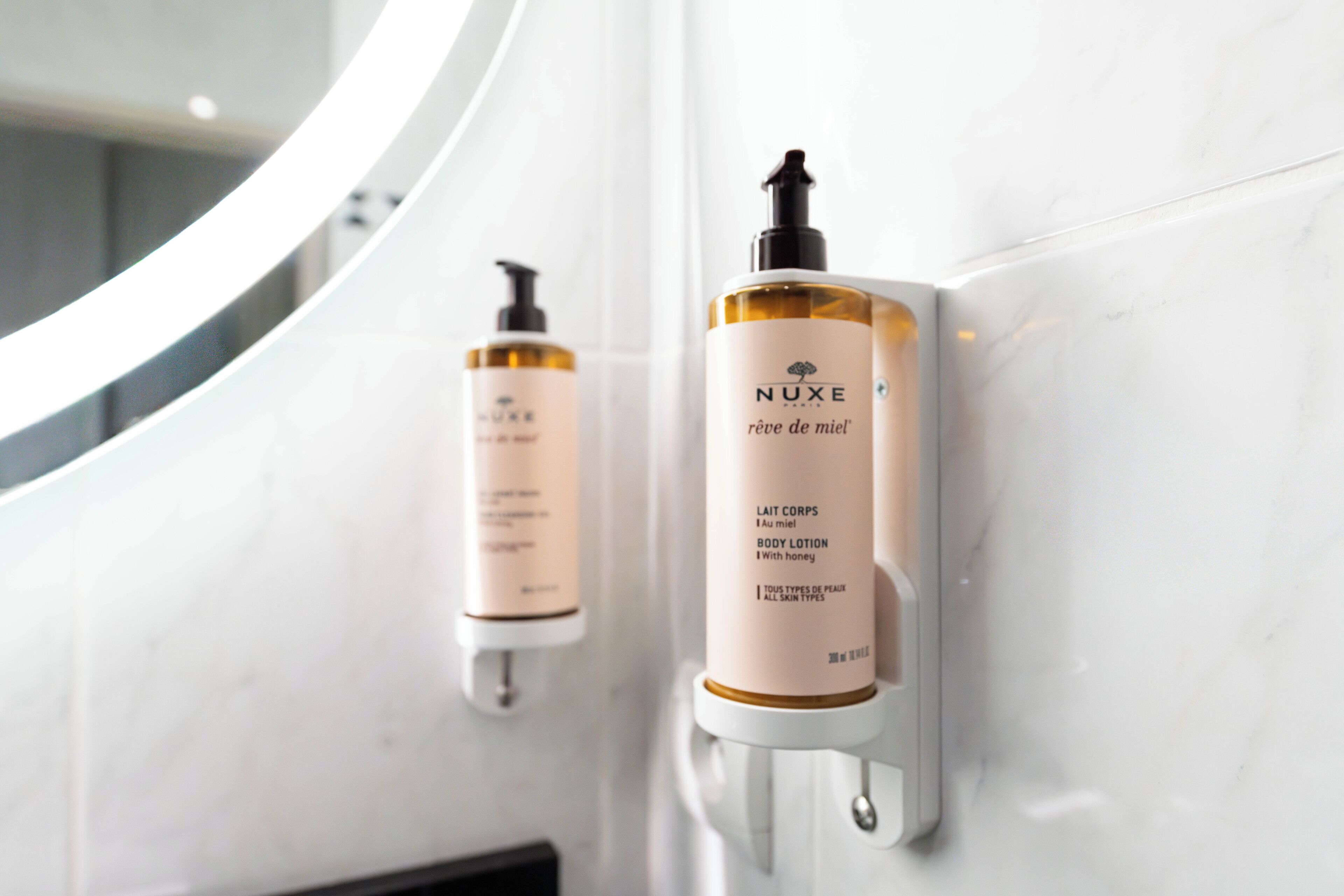 Two NUXE body lotion bottles mounted on a white marble wall.