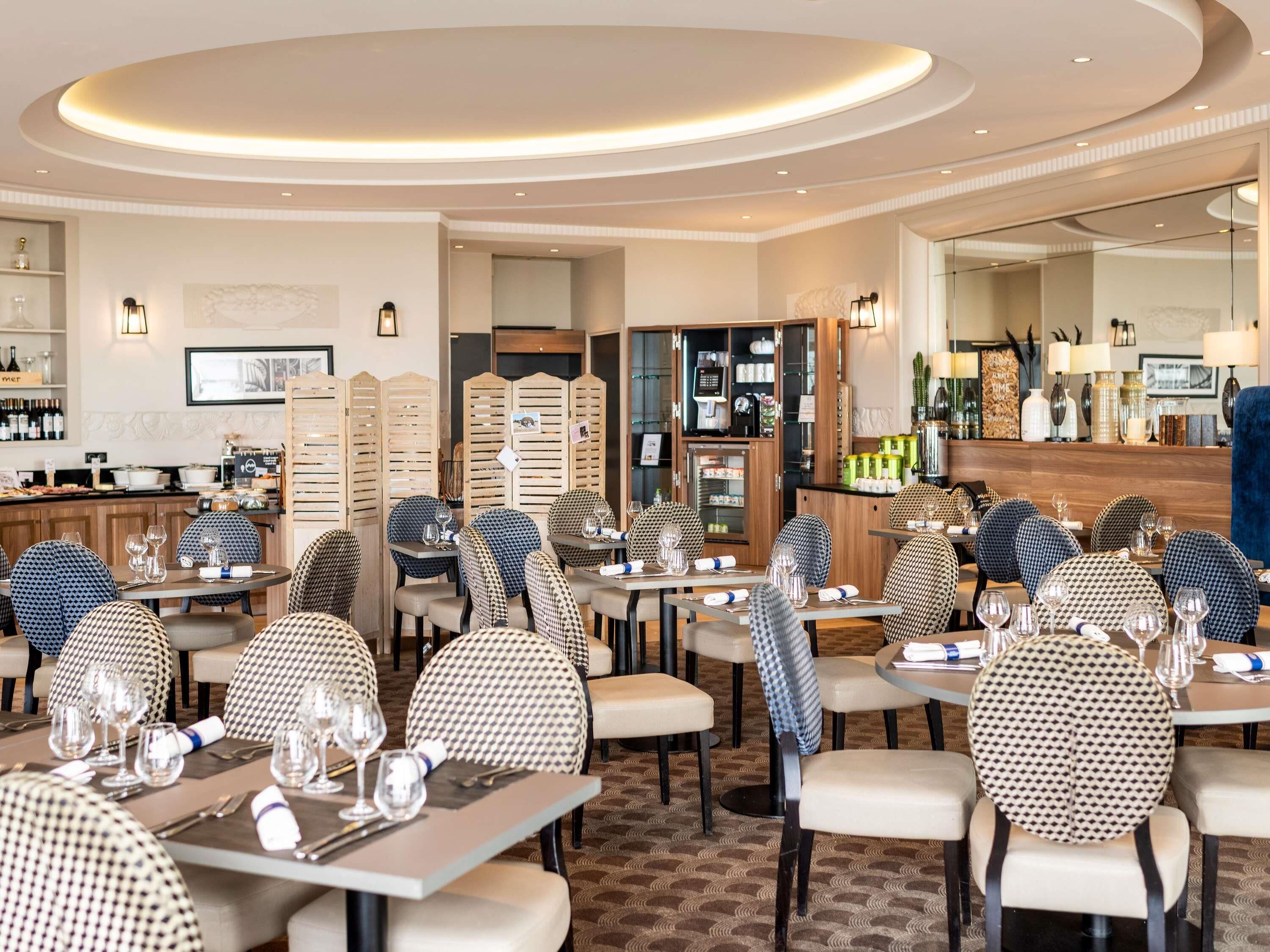 A restaurant with round tables, houndstooth patterned chairs, and a bar area with shelves of bottles.