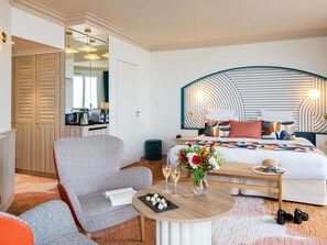 Une chambre d’hôtel moderne avec un grand lit, un coin salon comprenant une table, une chaise et une petite table à manger agrémentée d’un vase de fleurs et de quelques collations.