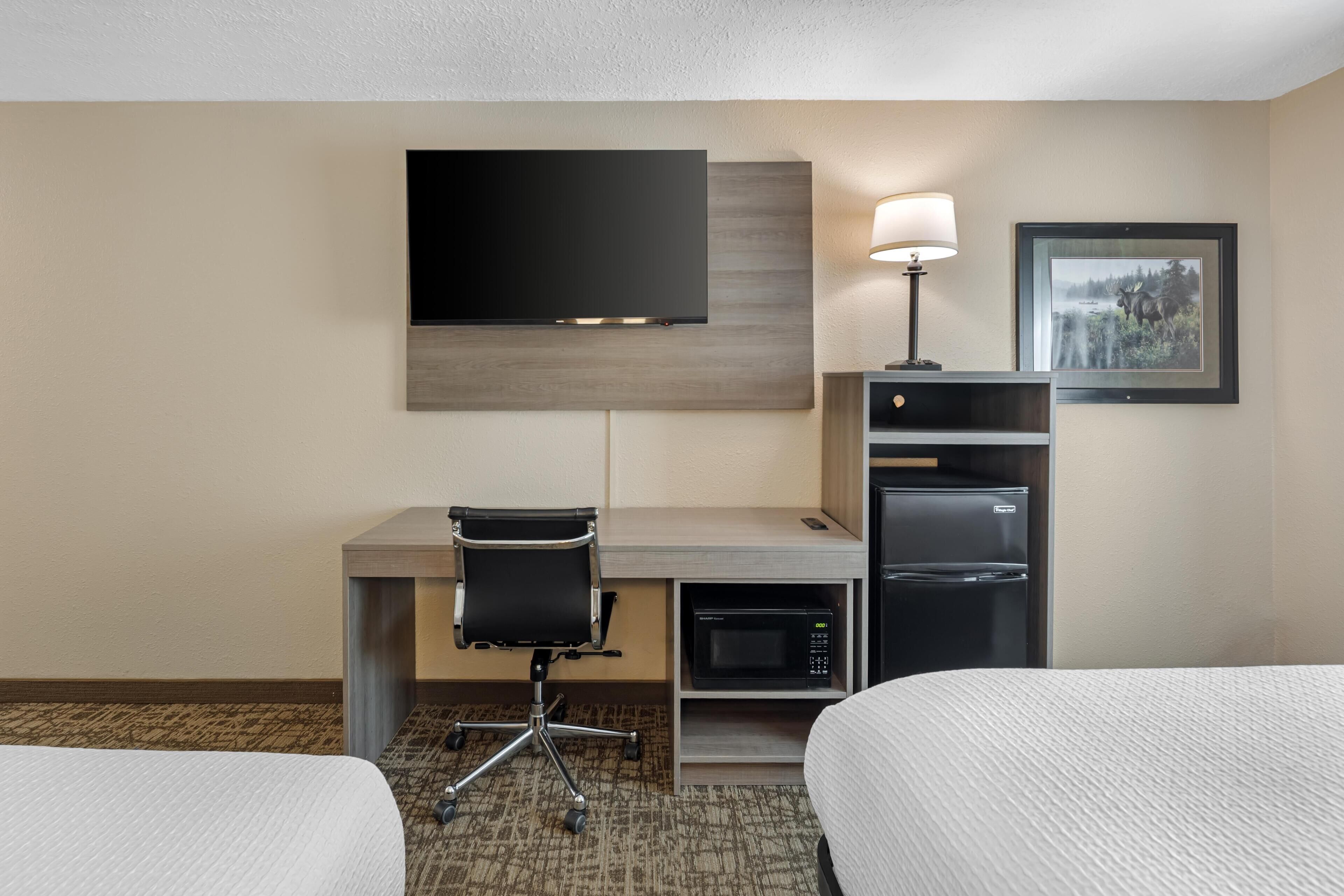 A hotel room with a desk, a flat-screen TV, a refrigerator, a lamp, and two beds.