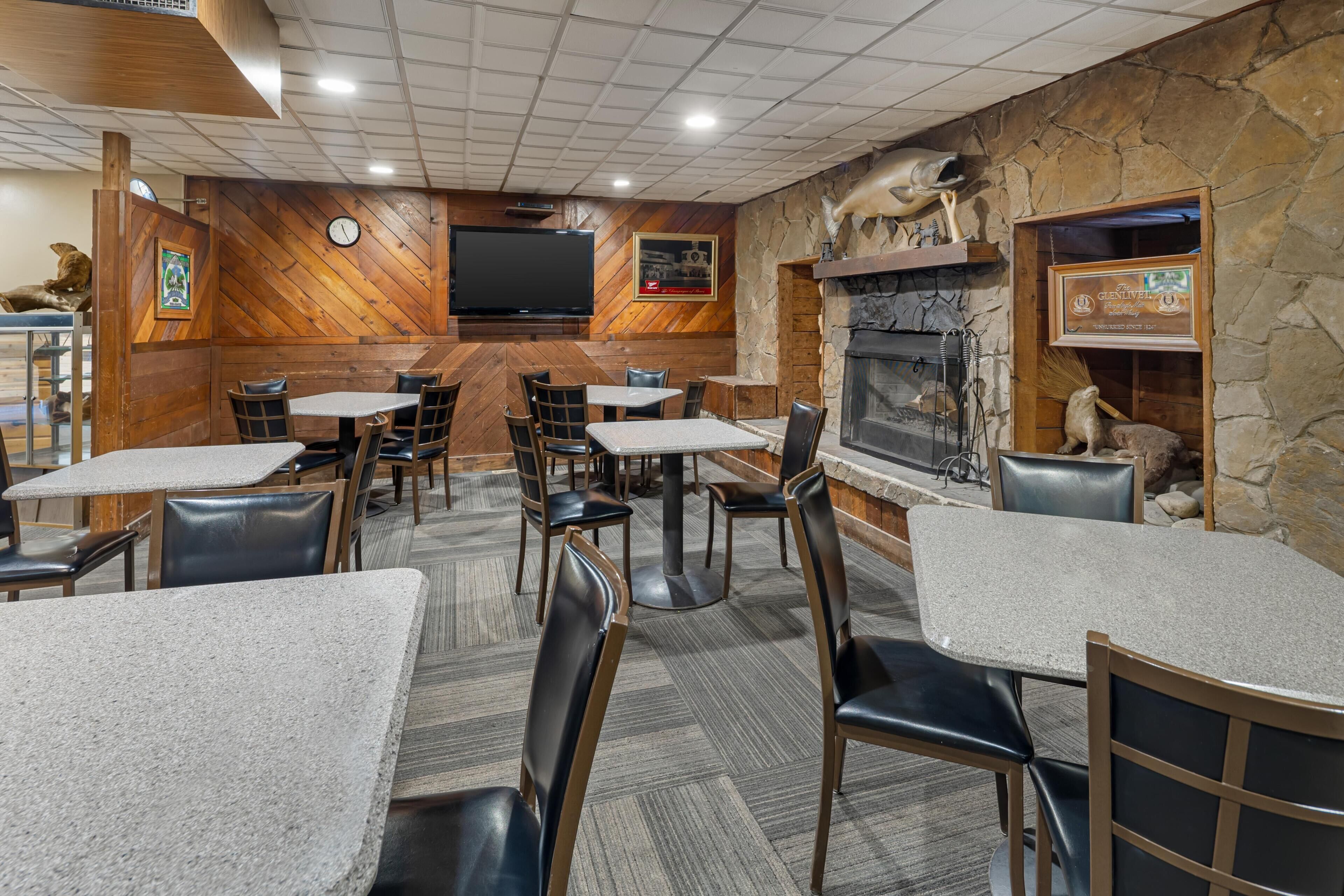 L’intérieur d’un restaurant présente des murs en bois, des éléments décoratifs en pierre et une cheminée. Des tables et des chaises sont disposées pour accueillir les clients.