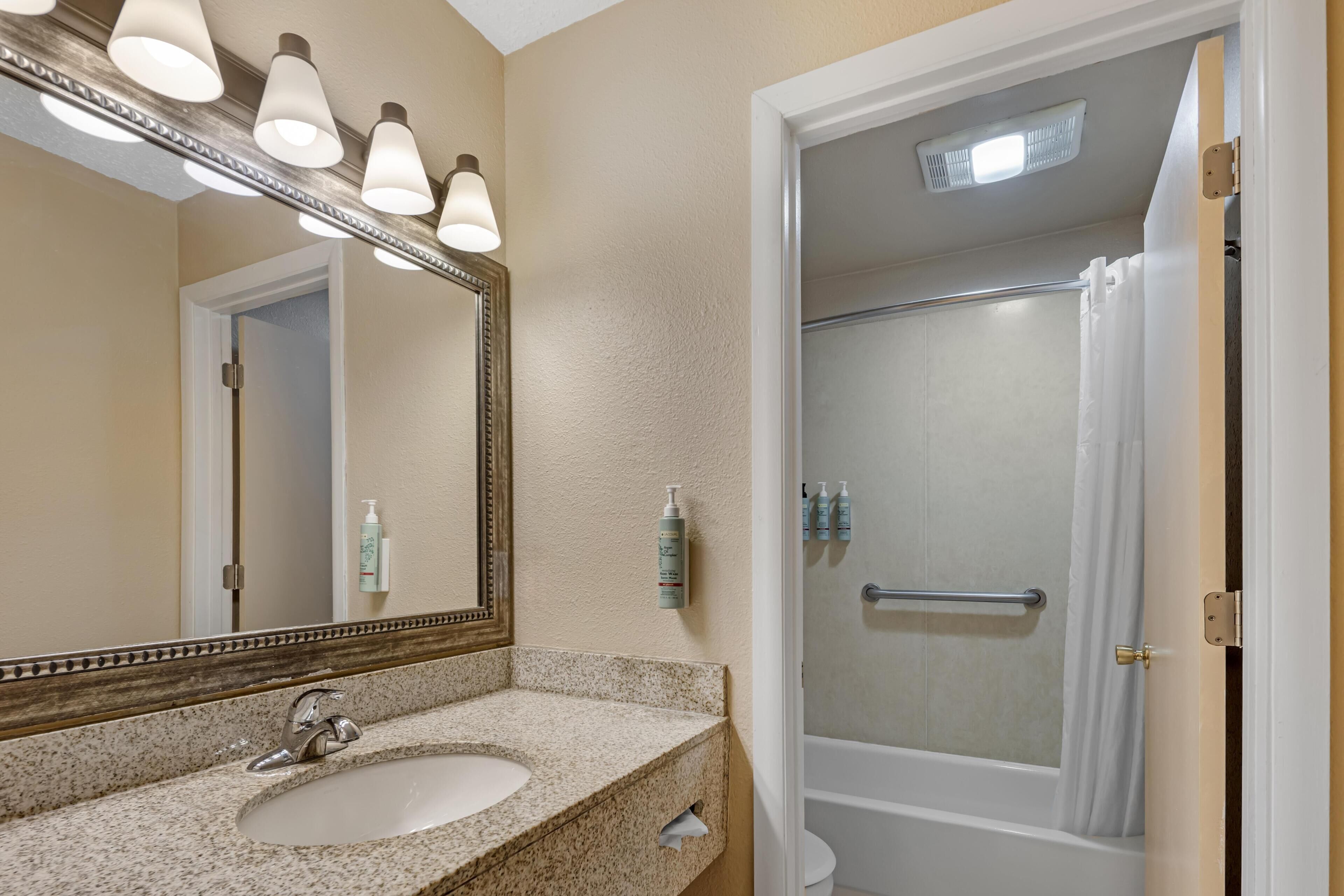 Un baño con lavamanos de encimera de granito, un espejo con iluminación, ducha con puerta de vidrio y artículos de aseo en la pared.