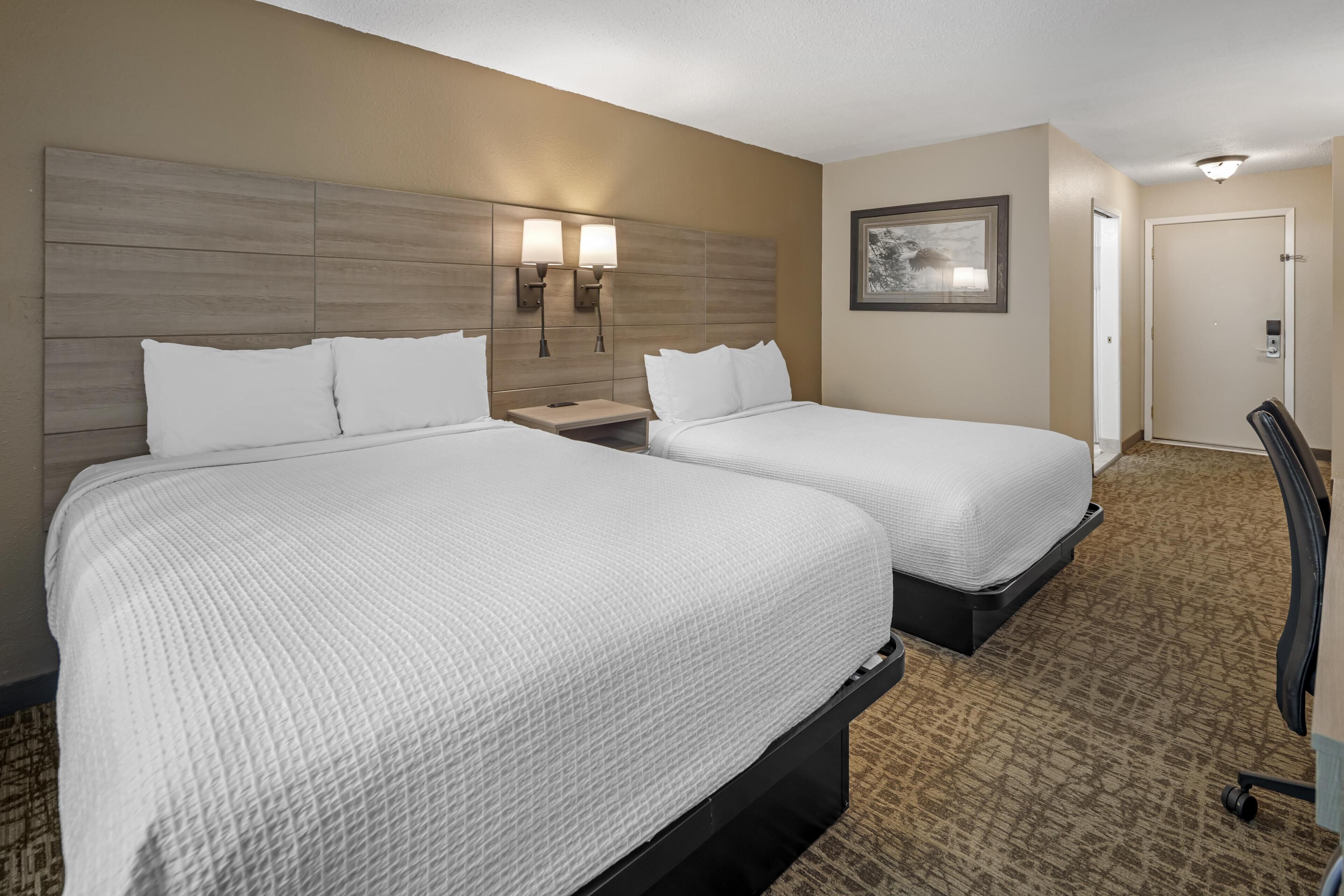 A hotel room with two beds, a nightstand, a wall-mounted lamp, a framed picture, and a door.
