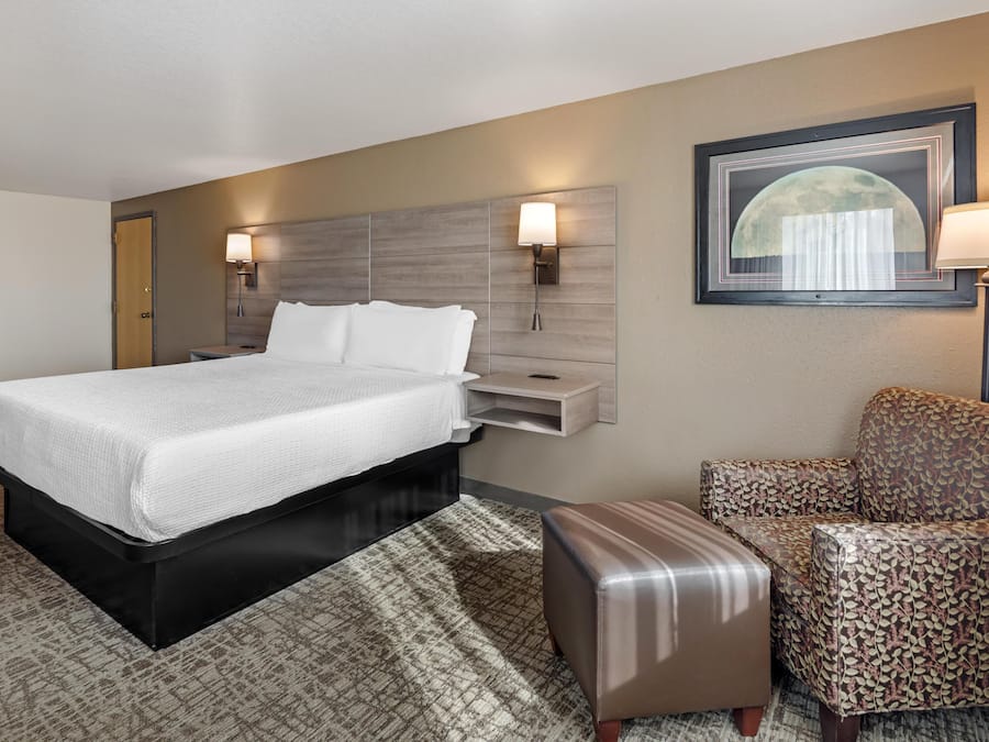 Quarto de hotel com cama, mesas de cabeceira, uma cadeira e luminária na parede.
