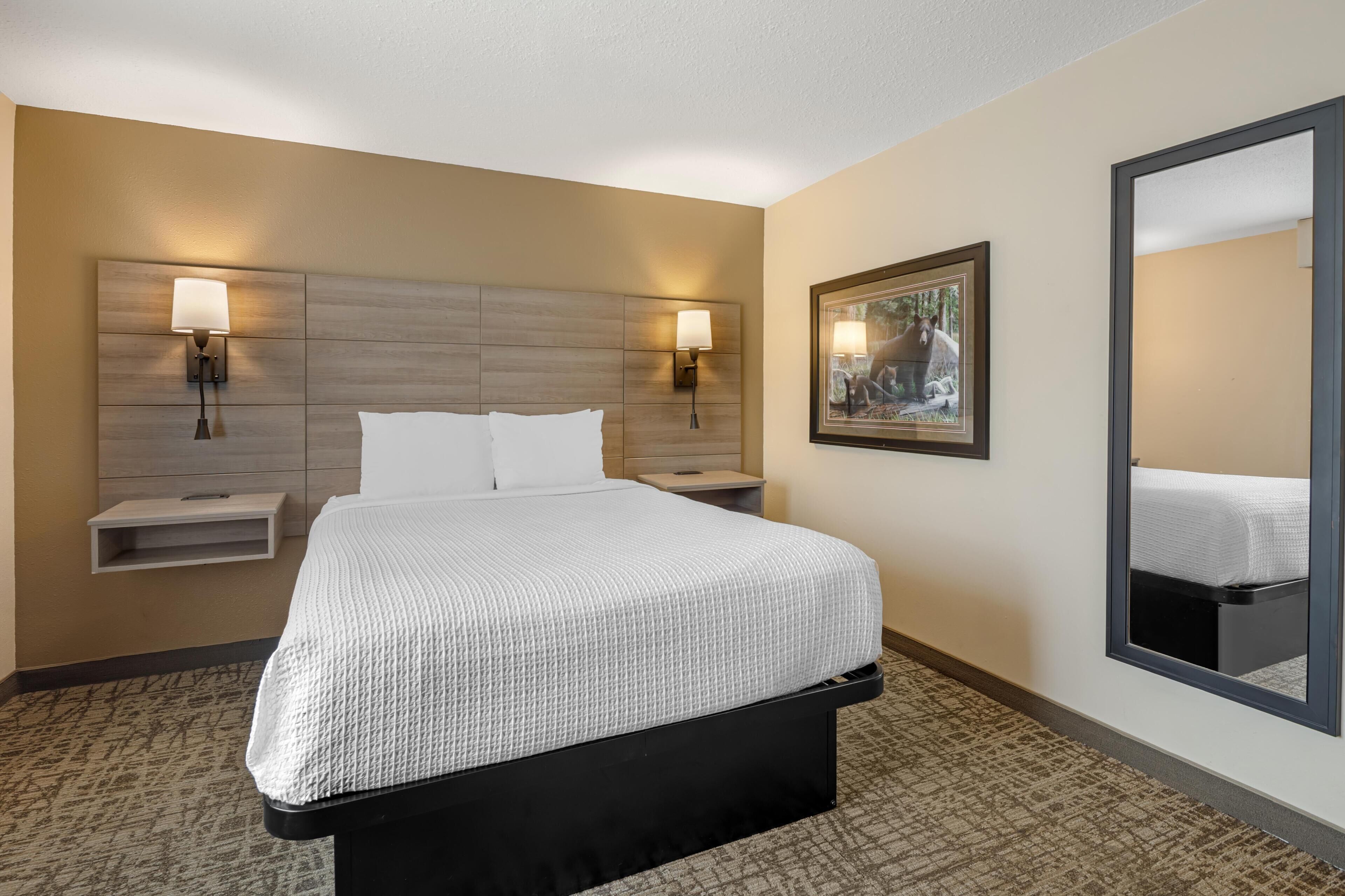 Habitación de hotel con una cama, dos mesitas de noche, una lámpara de pared y un cuadro enmarcado.
