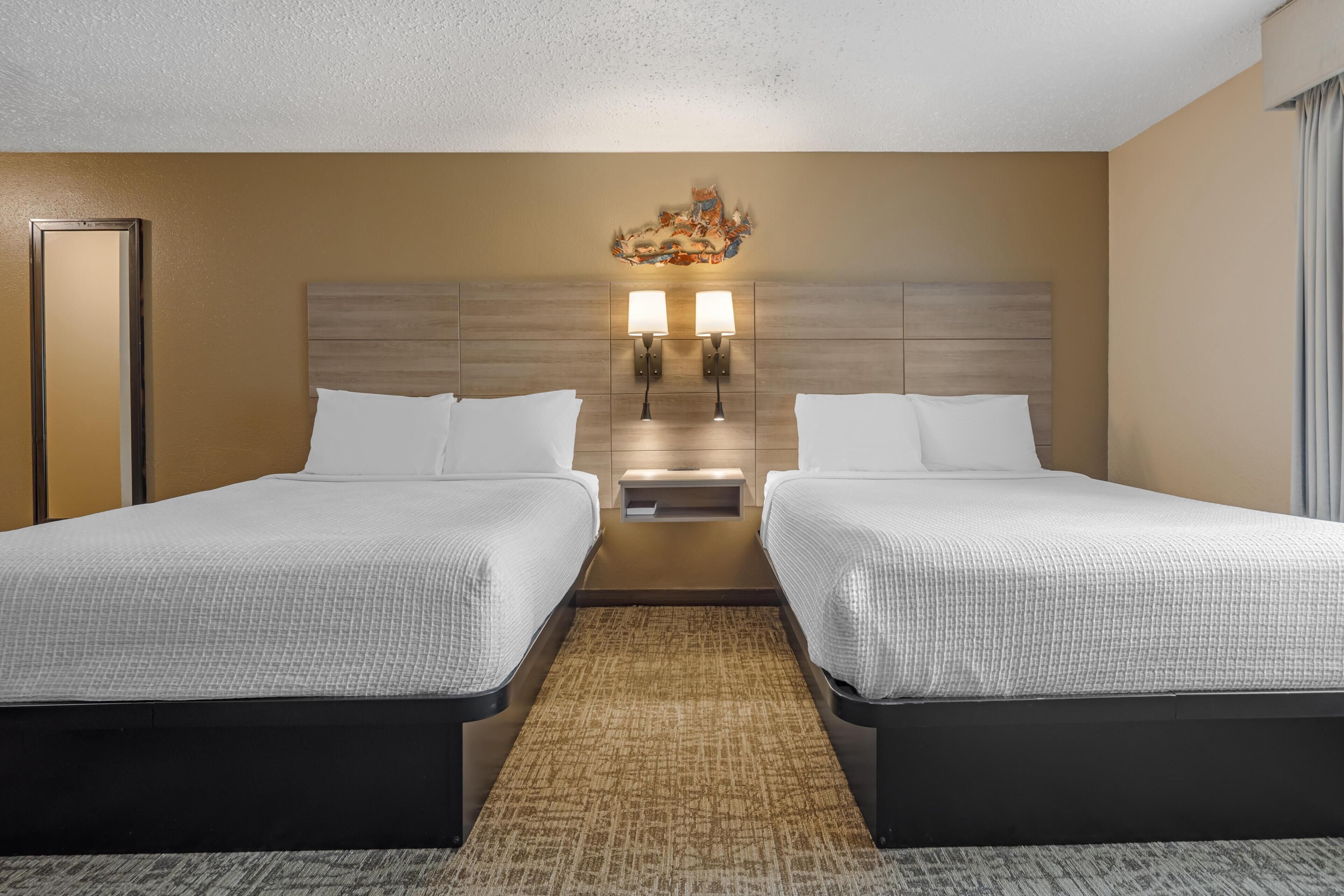 Ein Hotelzimmer mit zwei Betten, einem Kopfteil mit einer Leuchte und einem Teppichboden.