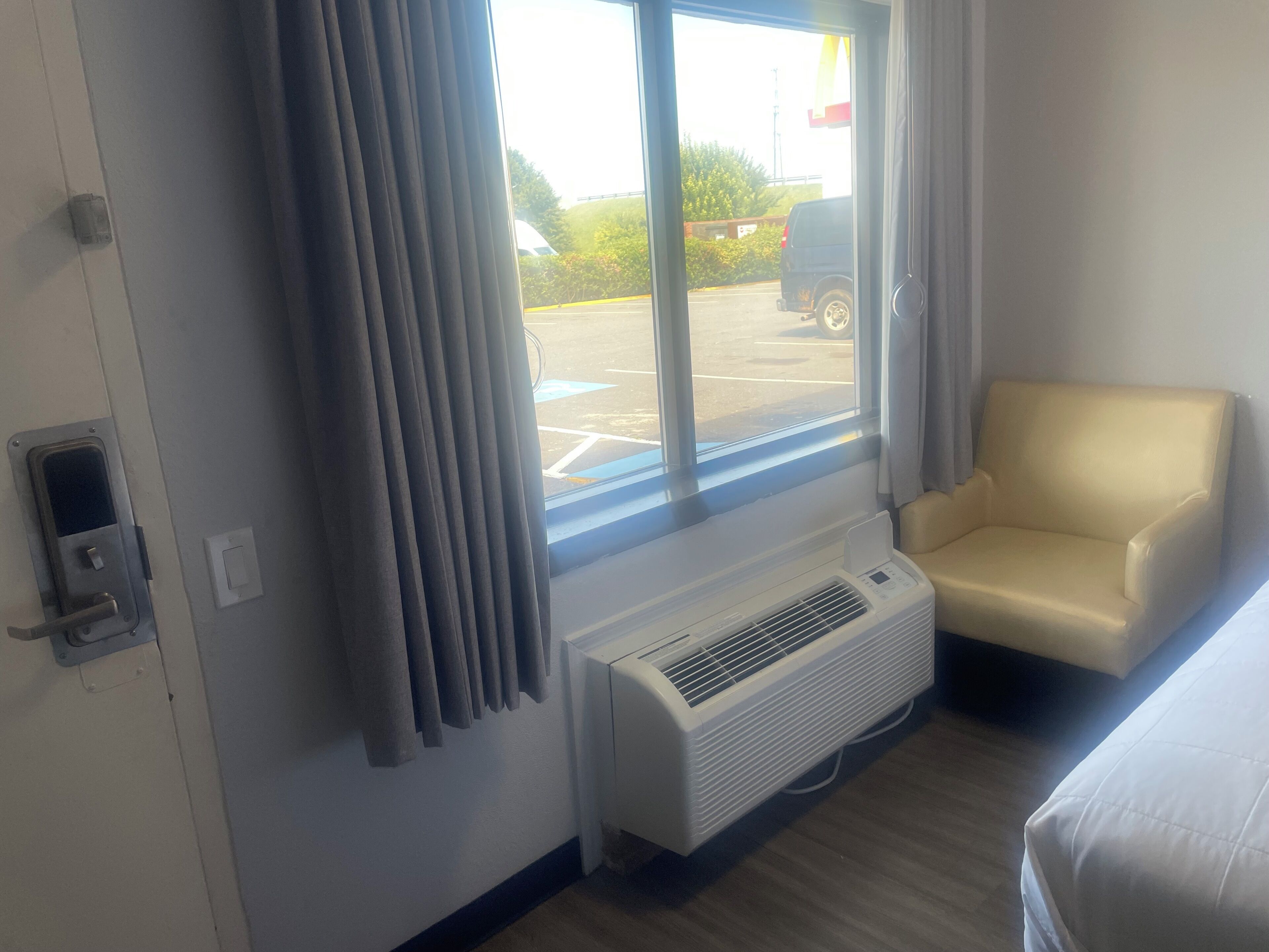 A hotel room with a large window, a white air conditioning unit, a beige armchair, and a door with a lock.