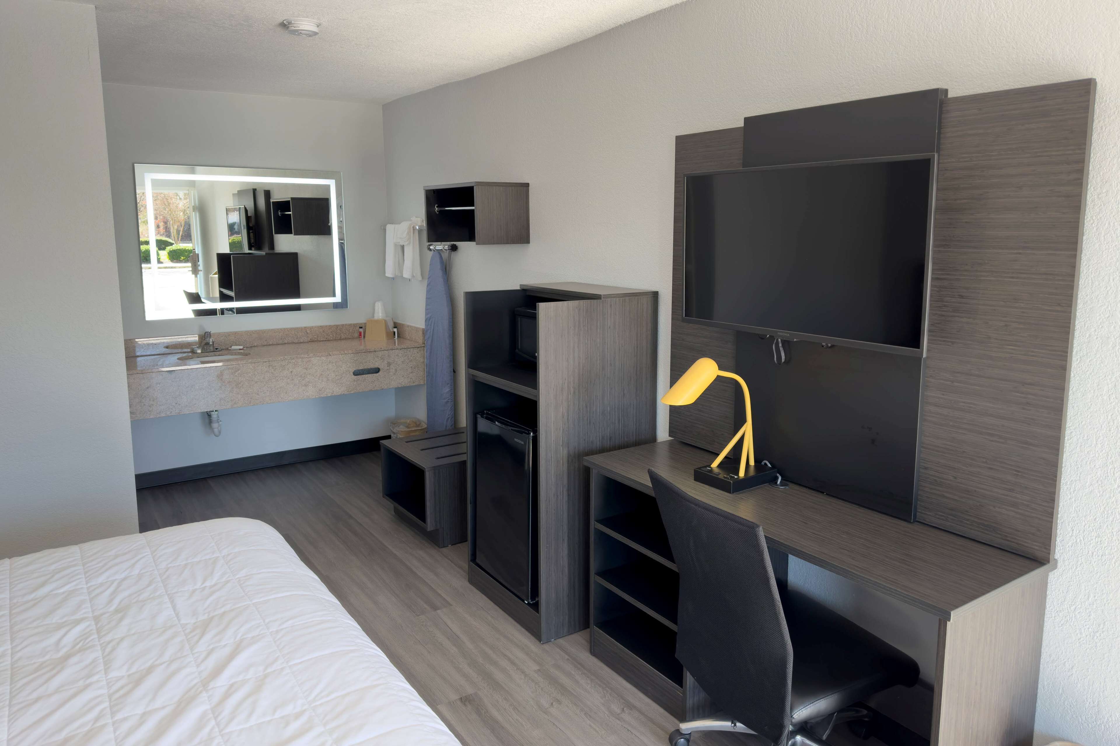 A modern hotel room with a bed, a desk with a chair, a television, a mirror, and a small kitchenette.