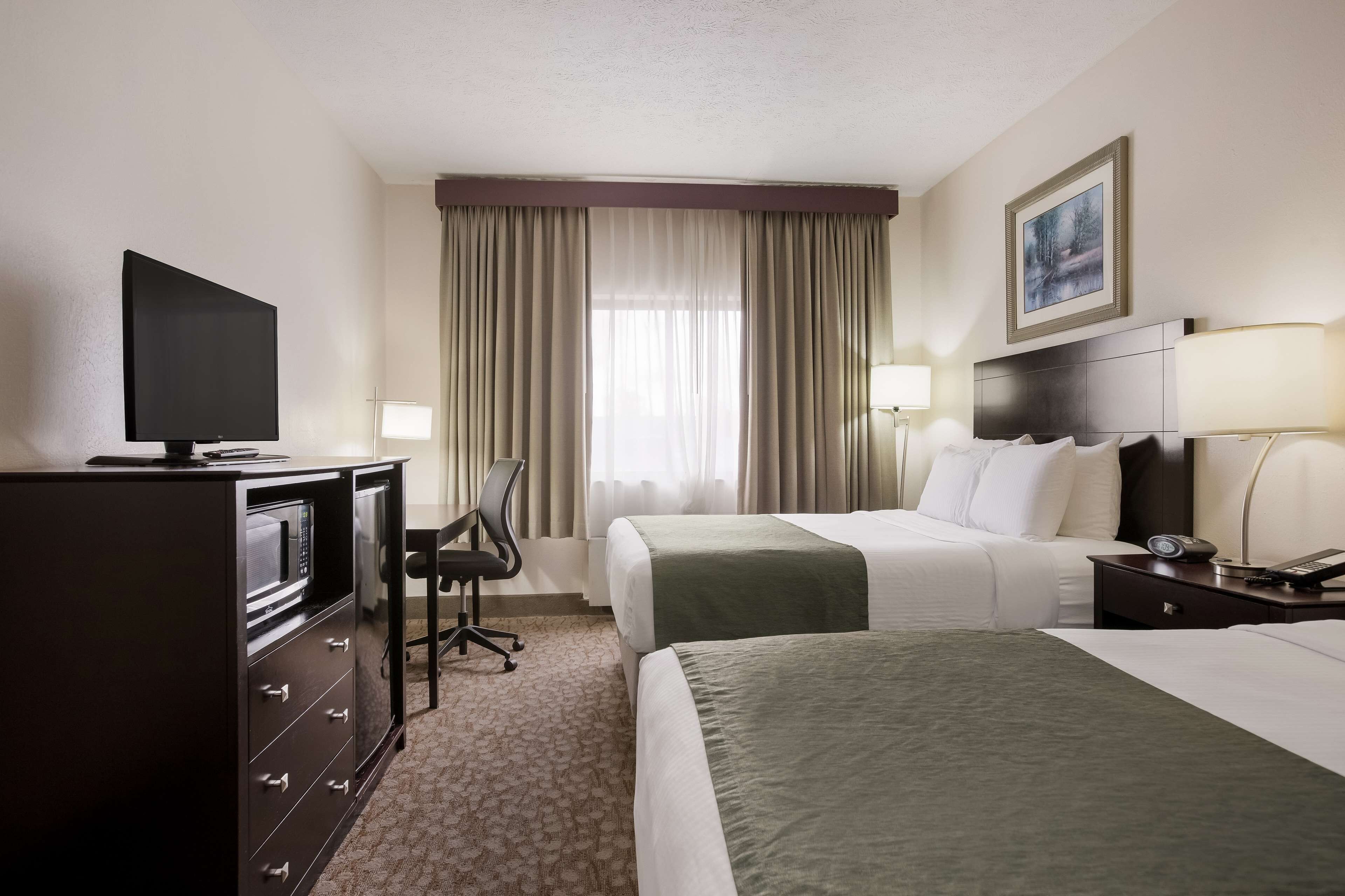 A hotel room with a bed, a desk with a TV, a chair, a lamp, and a painting on the wall.