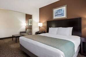 Habitació d'hotel amb un llit gran, una taula de nit, una cadira, una petita taula amb una làmpada i un quadre al mur.