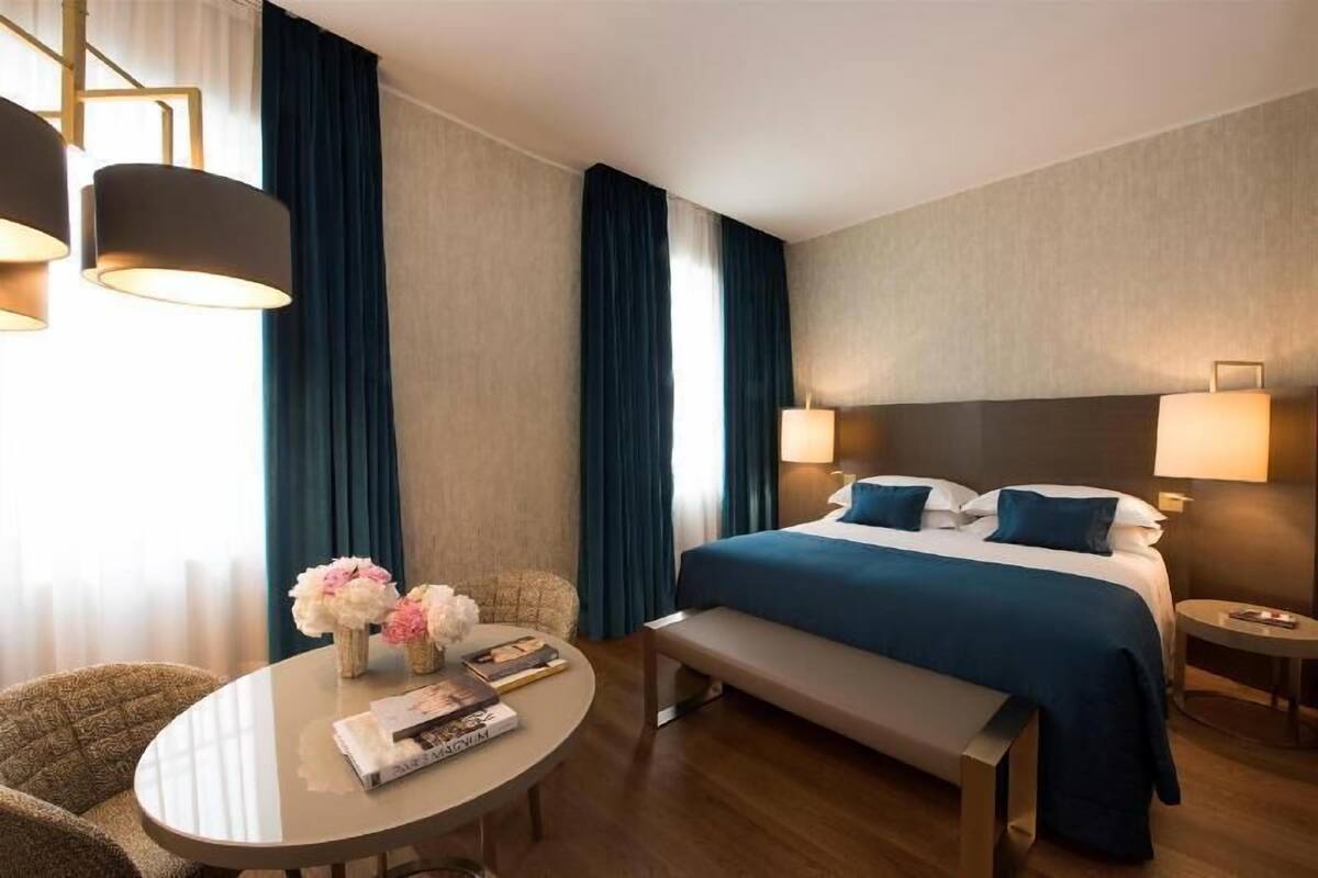 Une chambre d’hôtel avec un lit, des tables de chevet, une table ronde avec un banc et une fenêtre avec des rideaux.