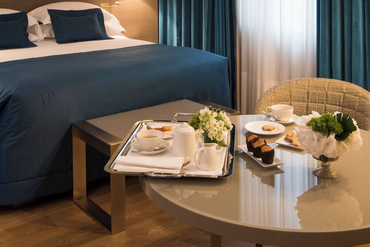 Une chambre d’hôtel avec un lit, une table de chevet sur laquelle se trouve un plateau avec des articles pour le petit-déjeuner, une lampe et une vue sur l’extérieur à travers les rideaux.