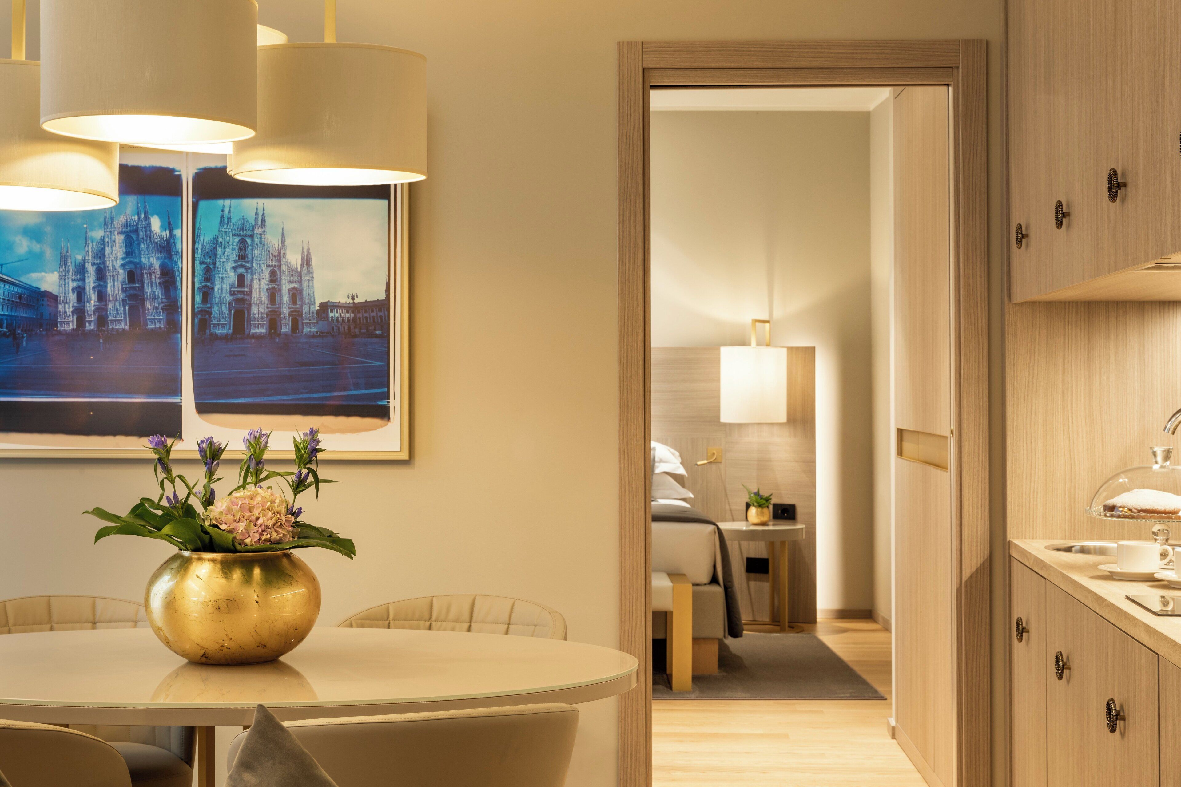 Habitación de hotel con zona de comedor que incluye una mesa redonda, sillas y un jarrón con flores. Se observa una cama con mesita de noche y lámpara, además de vista del cuarto a través de una puerta abierta.