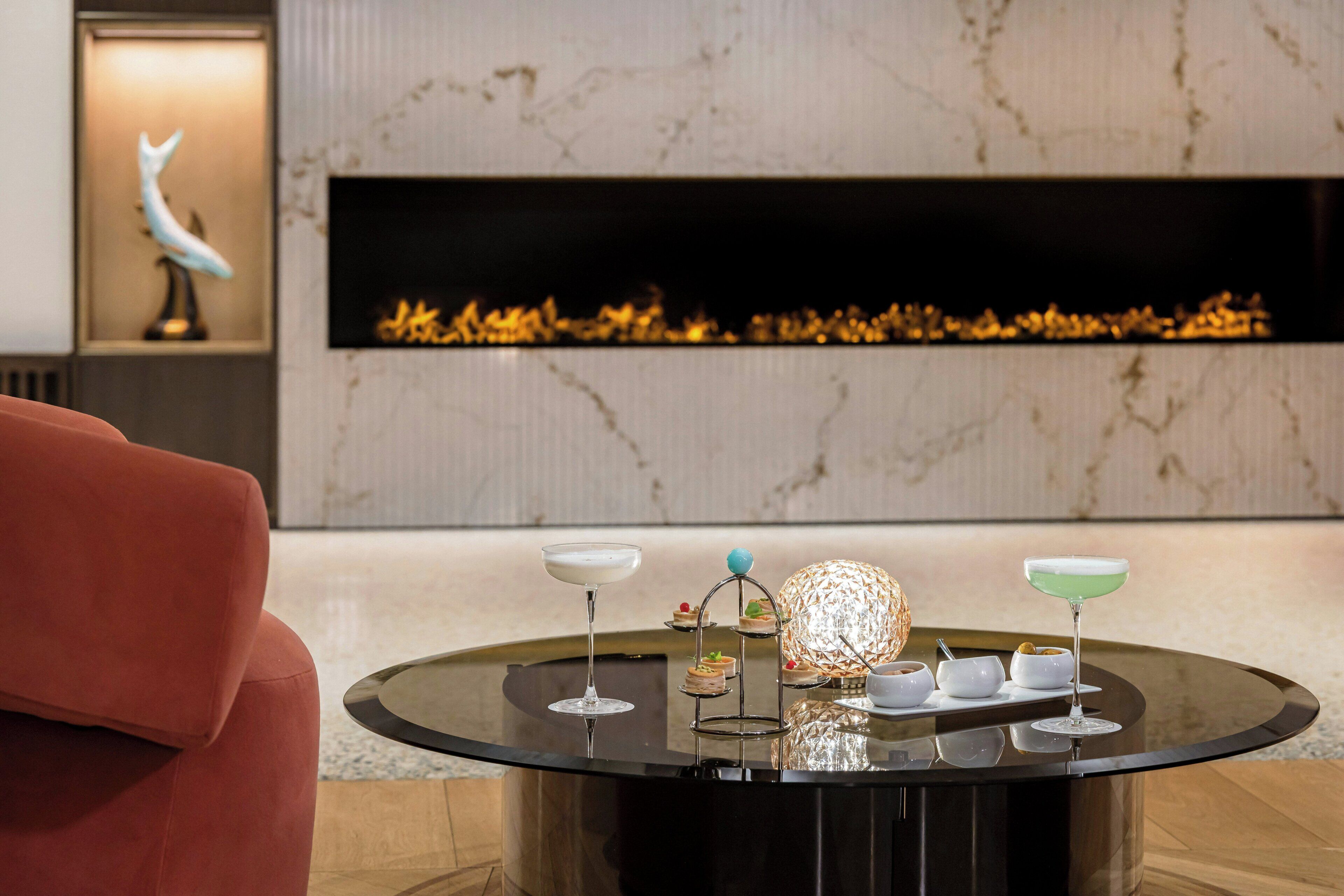 Een modern interieur met een ronde glazen tafel waarop diverse drankjes en desserts staan, een rode bank en een open haard met een decoratieve beeldhouwsel van een vogel.