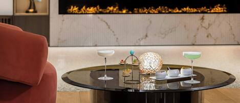 Een modern interieur met een ronde glazen tafel waarop diverse drankjes en desserts staan, een rode bank en een open haard met een decoratieve beeldhouwsel van een vogel.