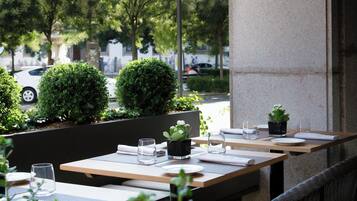 An outdoor seating area with tables and chairs, surrounded by greenery and a view of the street.
