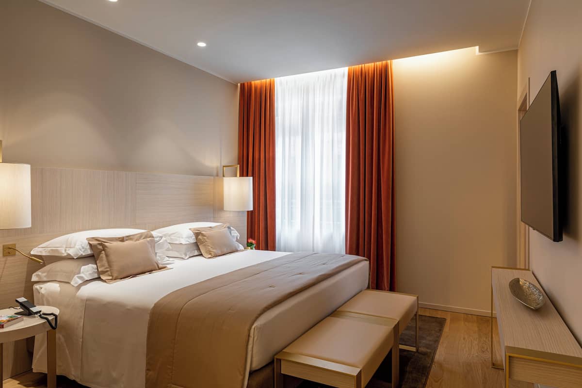 A modern hotel room with a large bed, bedside tables, a flat-screen TV, and a large window with curtains.