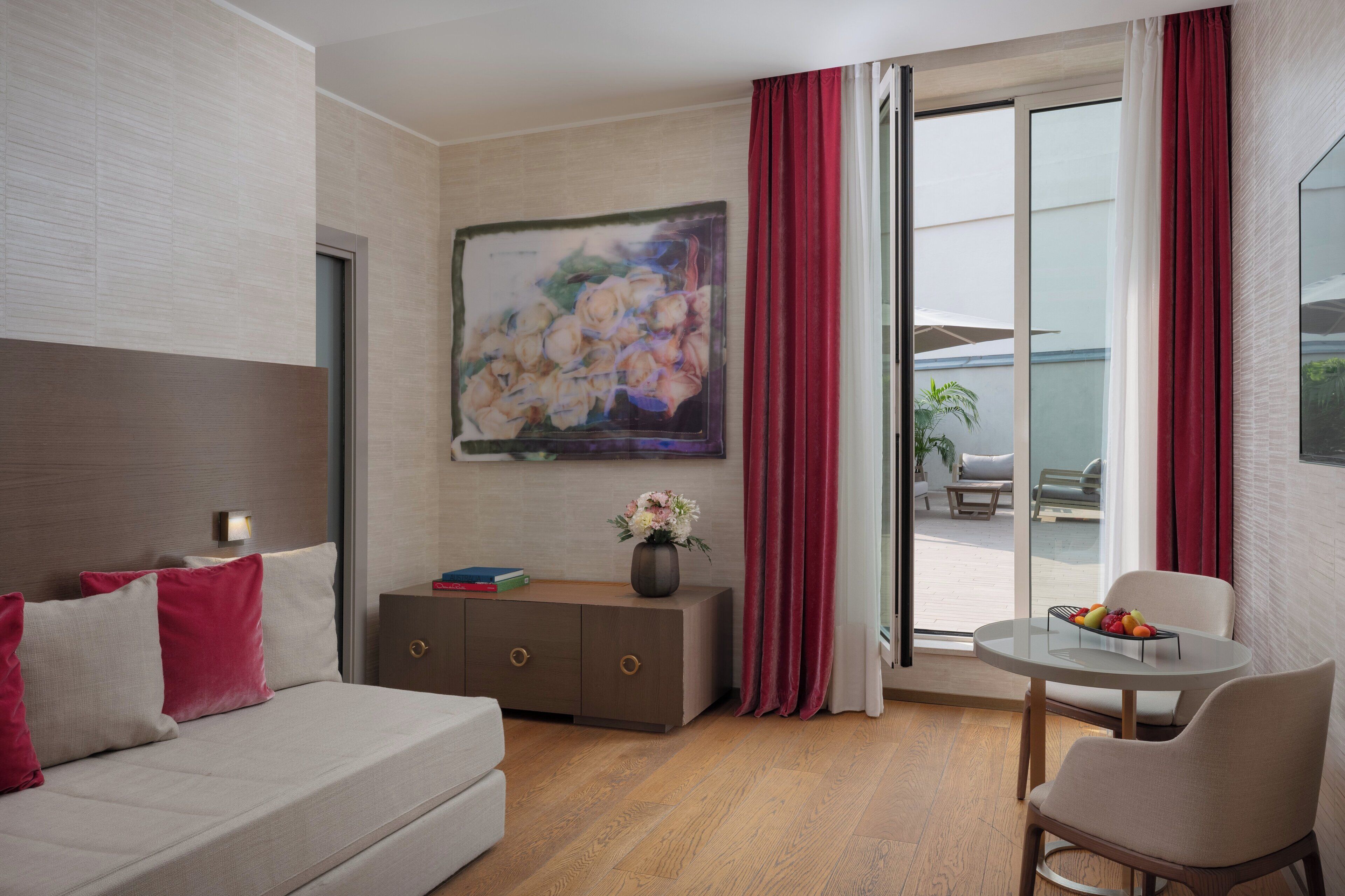 A modern hotel room with a sofa, a dining table with fruit, a vase with flowers, and a large painting on the wall.