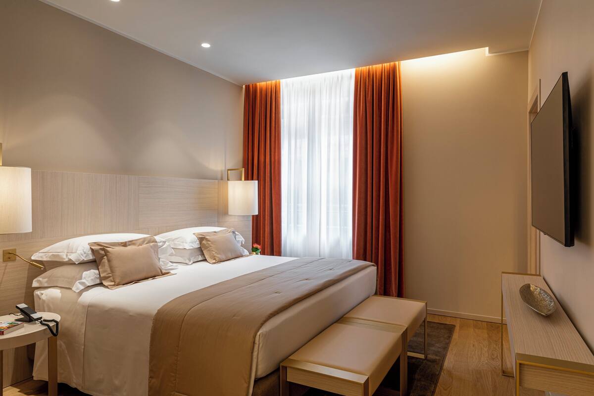 Une chambre d’hôtel moderne dotée d’un grand lit, de tables de chevet, d’un téléviseur à écran plat et d’une grande fenêtre avec des rideaux.