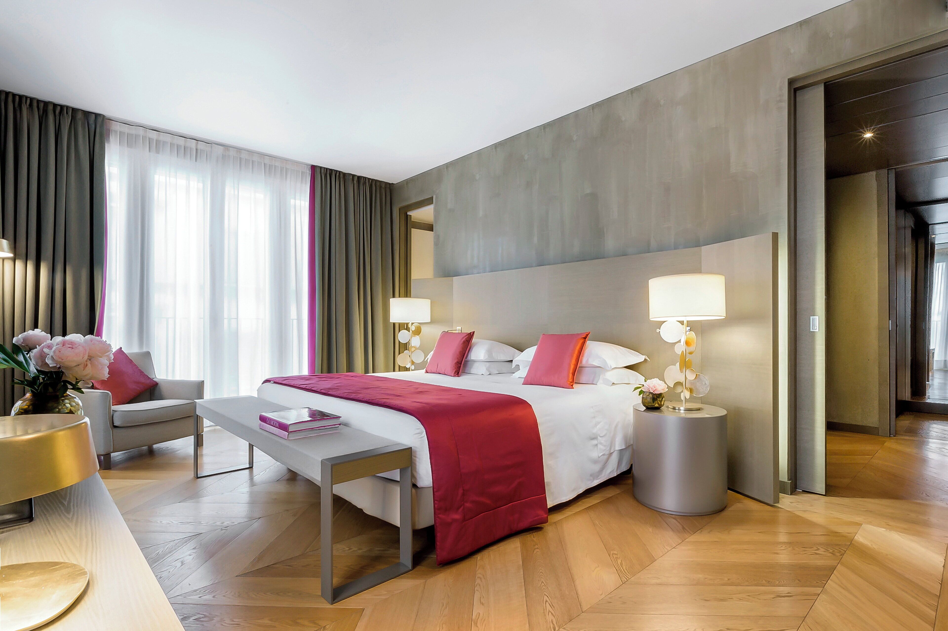 A modern hotel room with a large bed, two bedside tables with lamps, a bench, a desk, and a chair.
