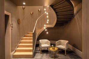 A modern interior with a wooden staircase, two white armchairs, a small round table, and a wall clock.