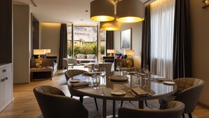 A modern dining area with a round table set for six, surrounded by comfortable chairs. There are two pendant lights above the table, and the room has a large window with curtains. - Rosa Grand Milano - Starhotels Collezione (Milan)