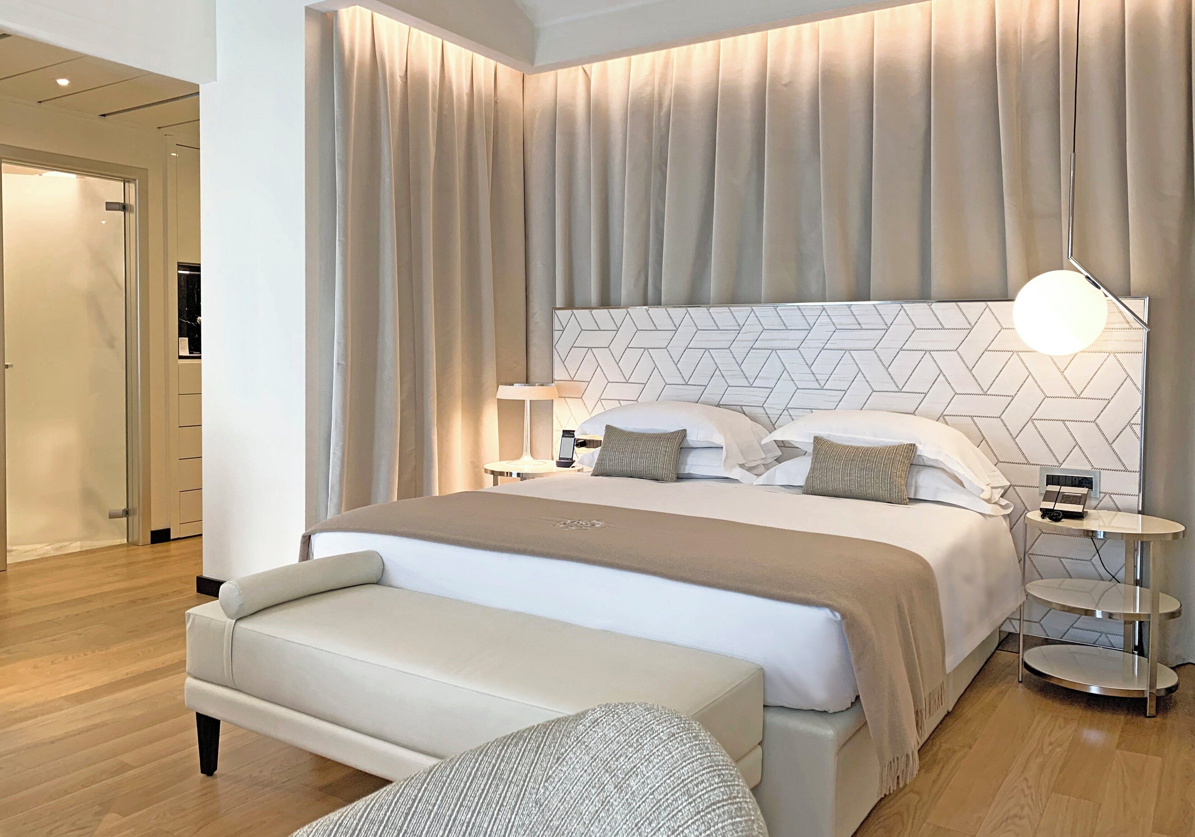 A modern hotel room with a large bed, a bedside table with a lamp, and a bench.