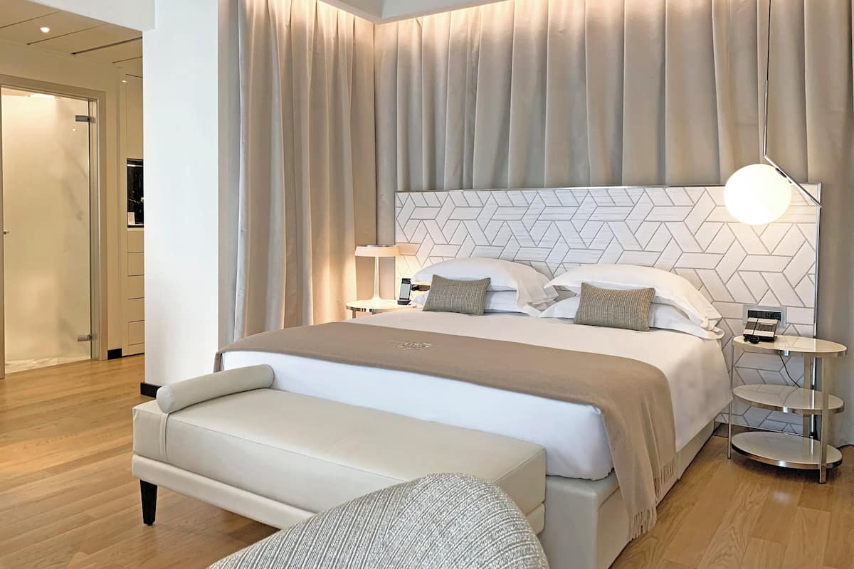 A modern hotel room with a large bed, a bedside table with a lamp, and a bench.