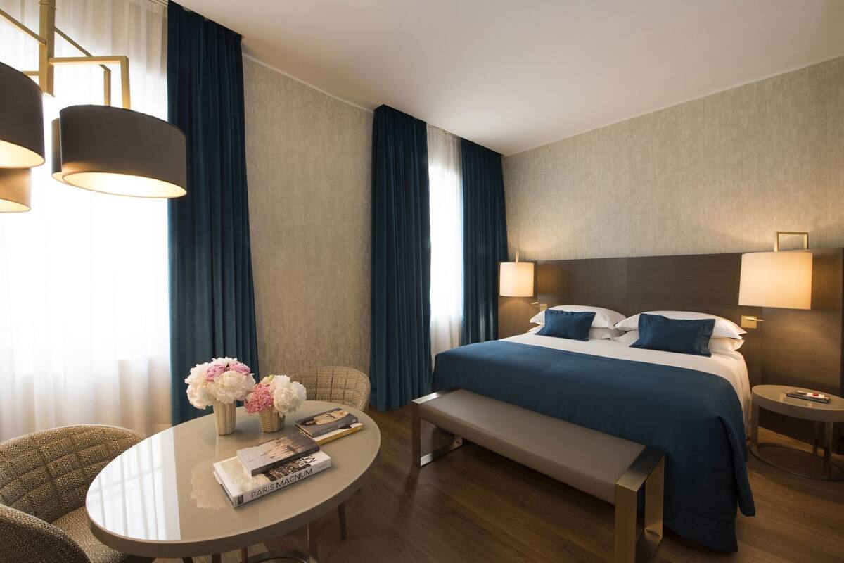 Une chambre d’hôtel comprenant un lit, des tables de chevet, une table ronde avec des chaises, un banc et un vase avec des fleurs.