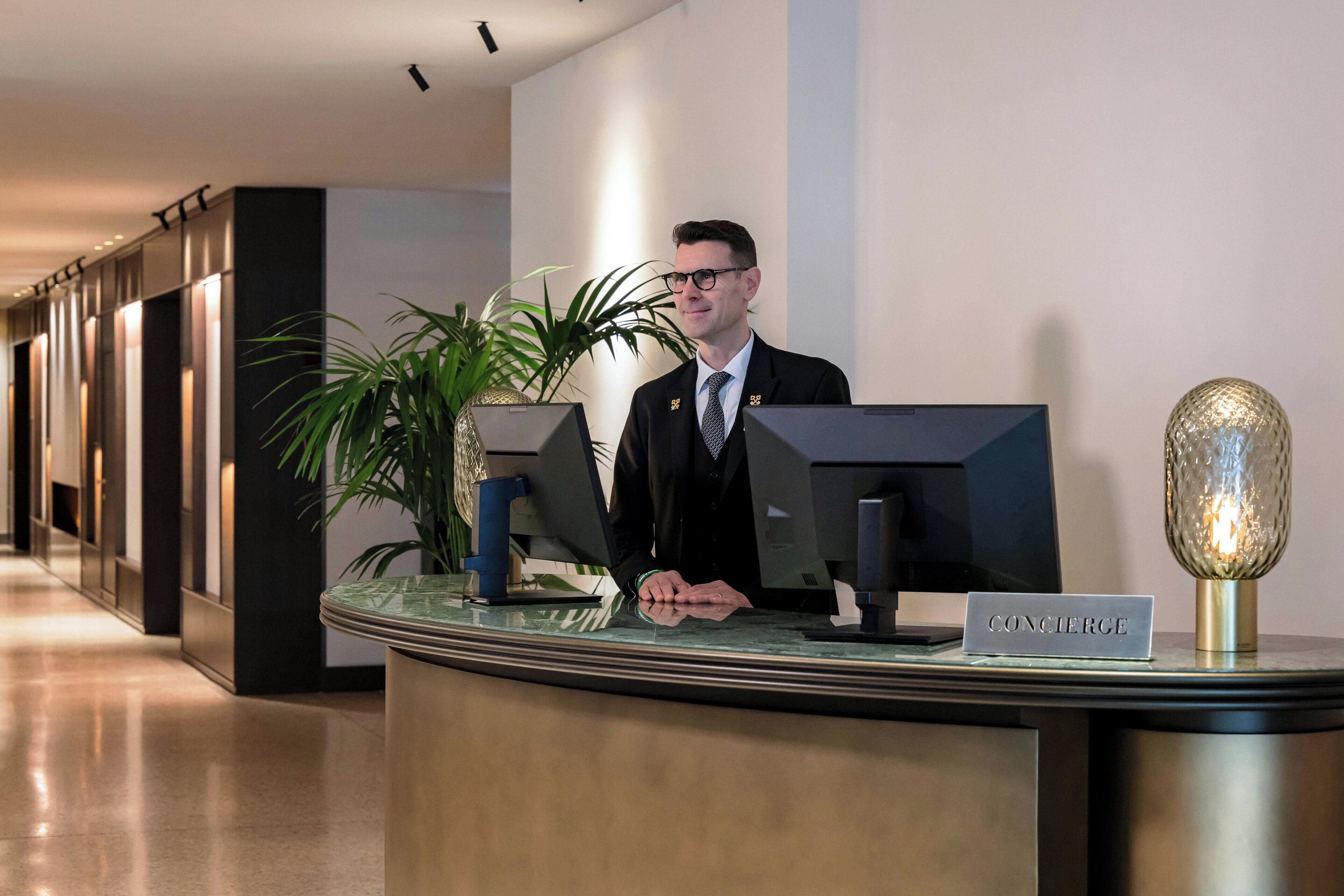 A reception desk with a 'CONCIERGE' sign, a computer monitor, a plant, and a decorative lamp.