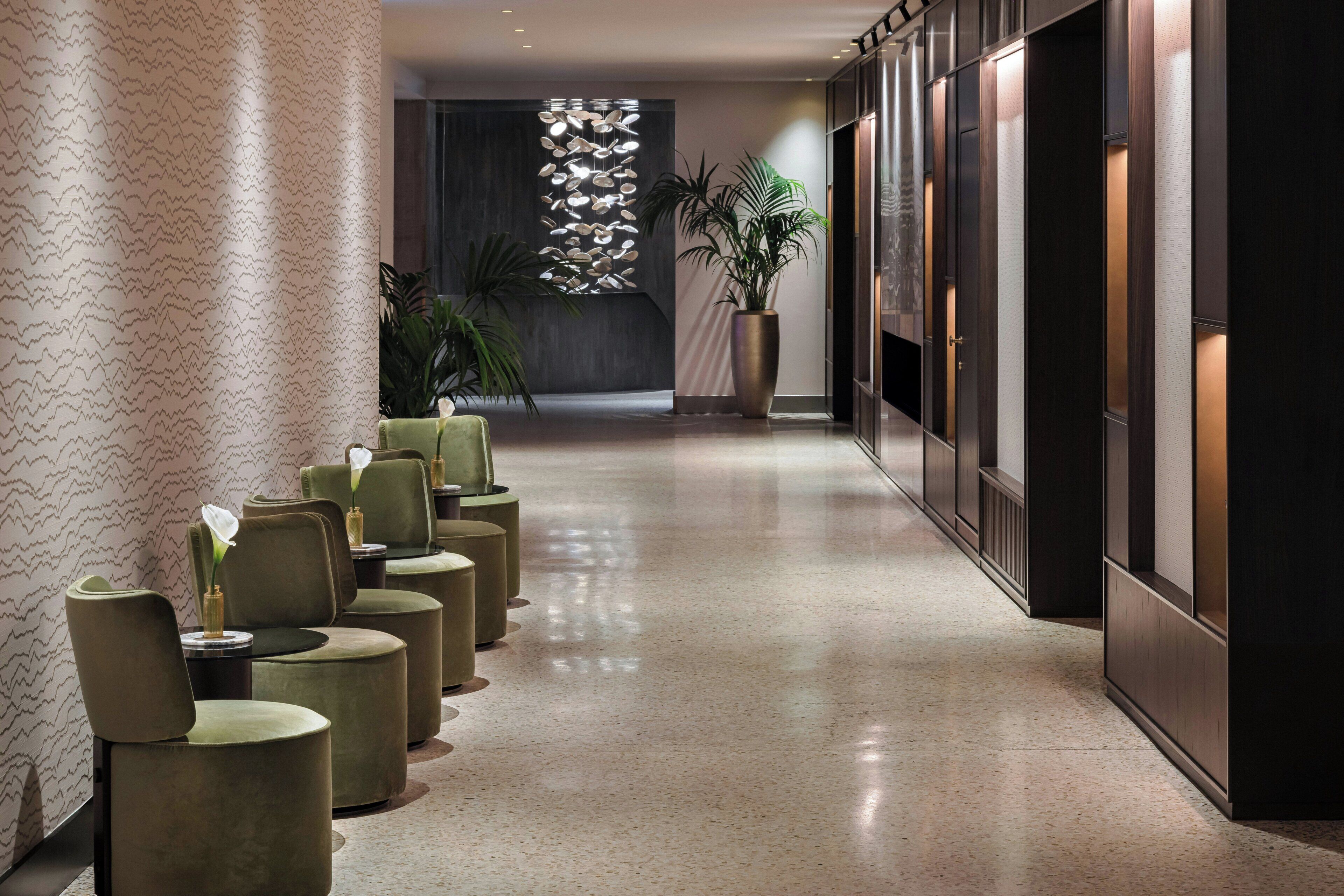 Un pasillo de hotel con una fila de sillas acolchadas verdes, una planta en maceta grande y una pared decorativa con un diseño geométrico.