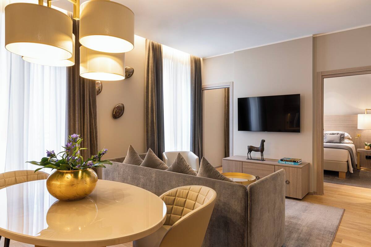 Une chambre d’hôtel moderne avec une table de salle à manger ronde, un canapé avec une chaise longue, un téléviseur fixé au mur et un lit avec une table de chevet.