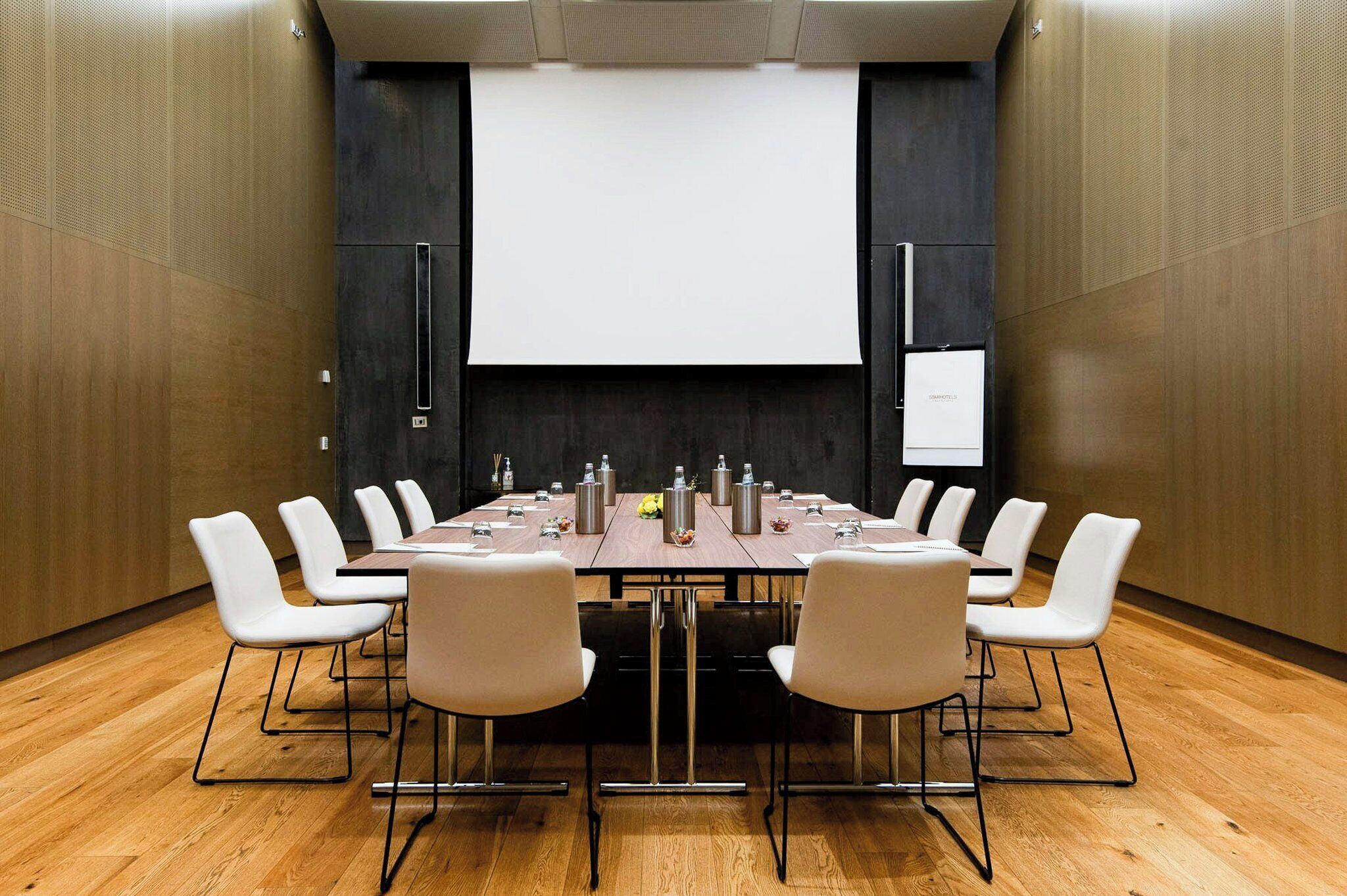 A conference room with a long table, white chairs, and a projector screen.