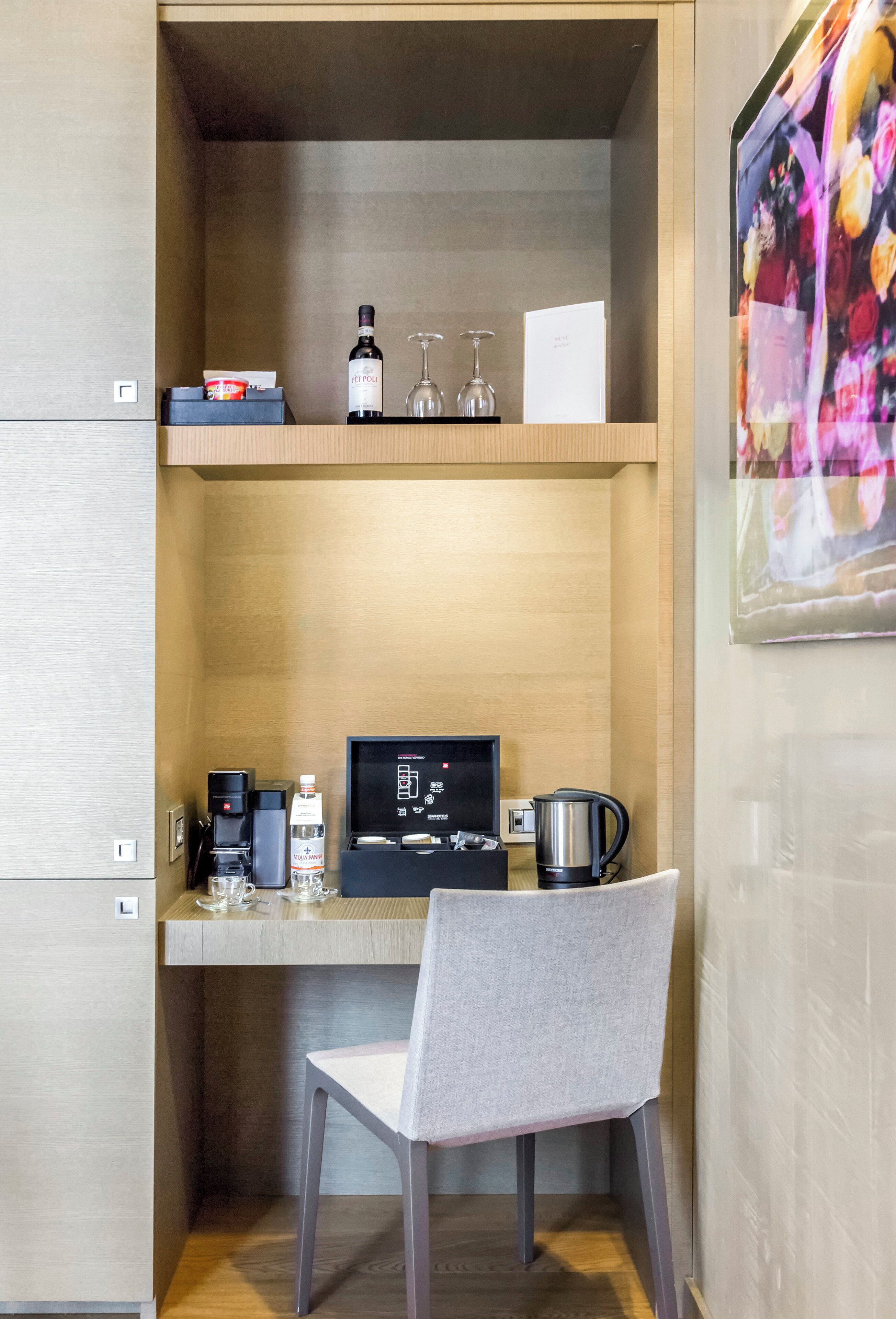 A compact hotel room with a wooden desk, a chair, a coffee maker, a bottle of water, a glass, and a small shelf with a bottle and a glass.