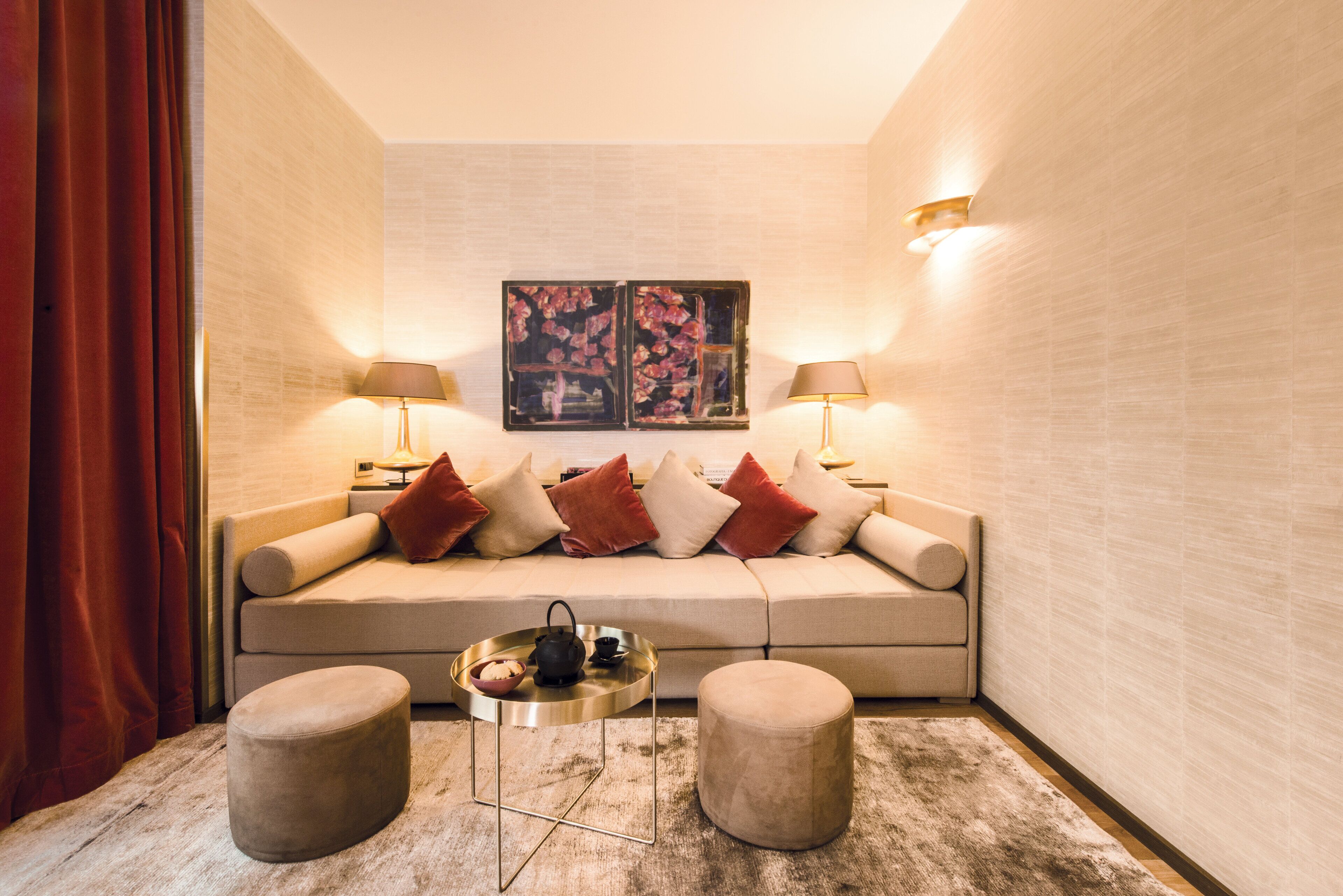 A cozy corner with a beige sofa, red and white cushions, two round ottomans, a small side table with a tray, and two wall-mounted lamps.