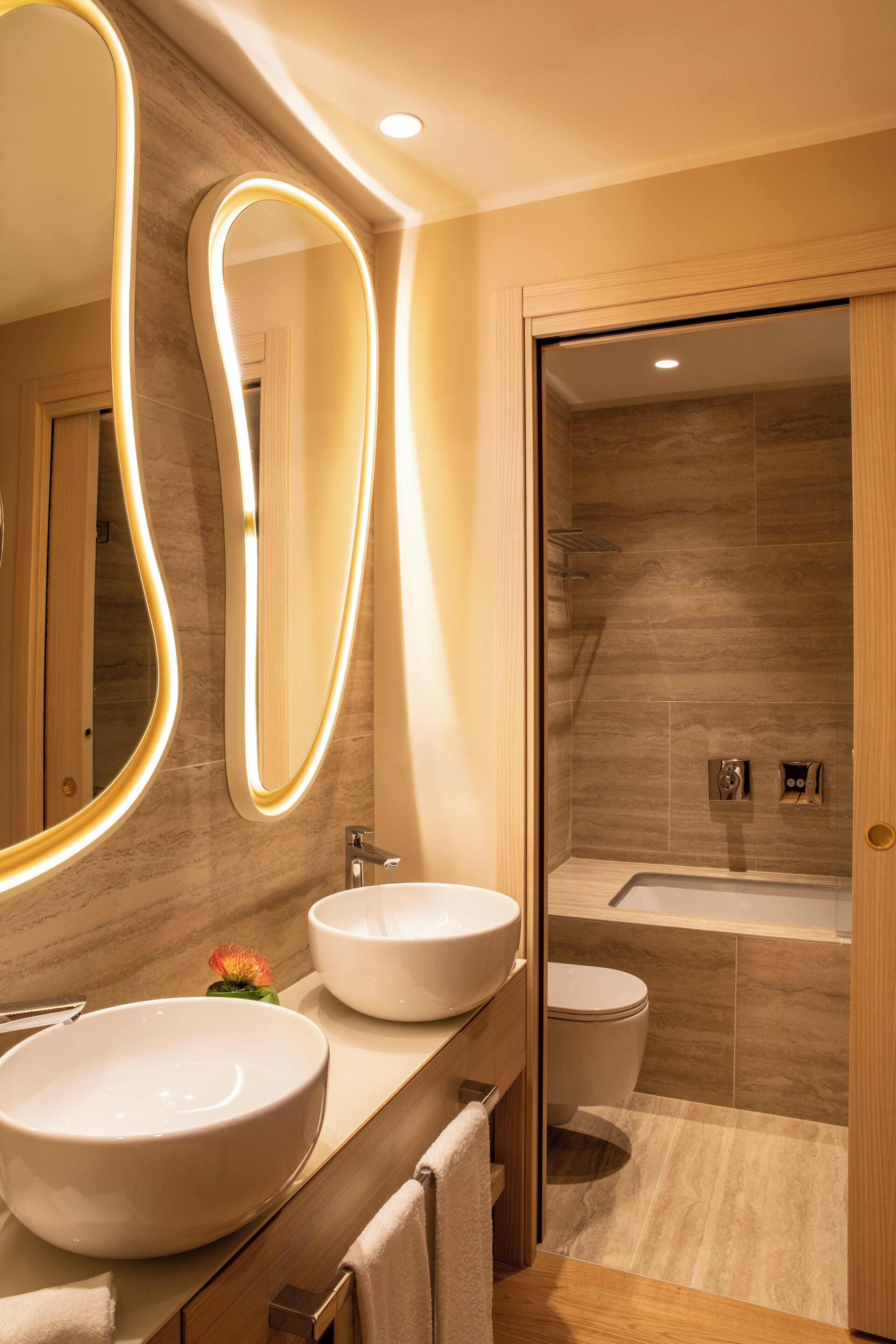A modern bathroom with two large sinks, a mirror with illuminated edges, a toilet, and a shower area.