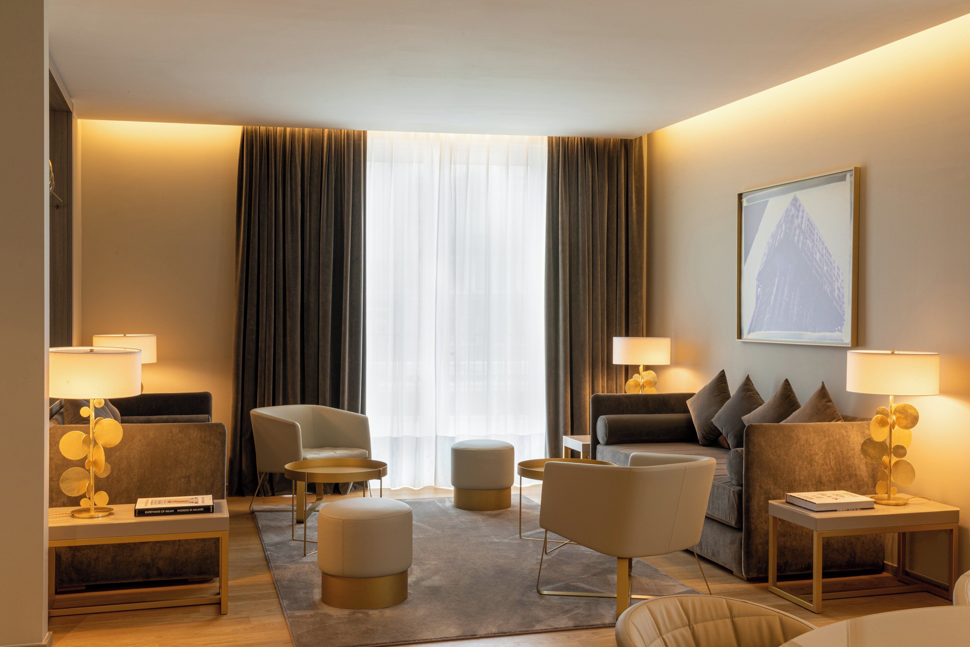 A modern hotel room with a sofa, armchairs, a coffee table, and a side table with a lamp.