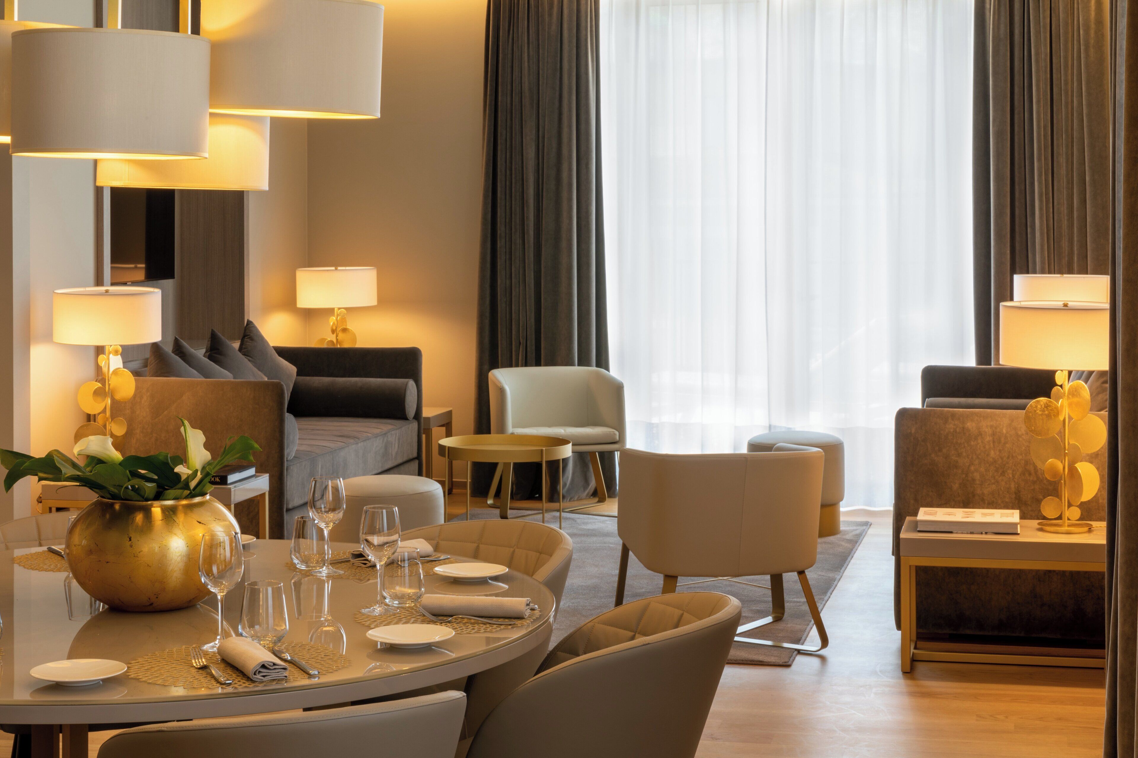 A hotel room with a round dining table set for four, a sofa, a chair, and a small side table with a lamp.