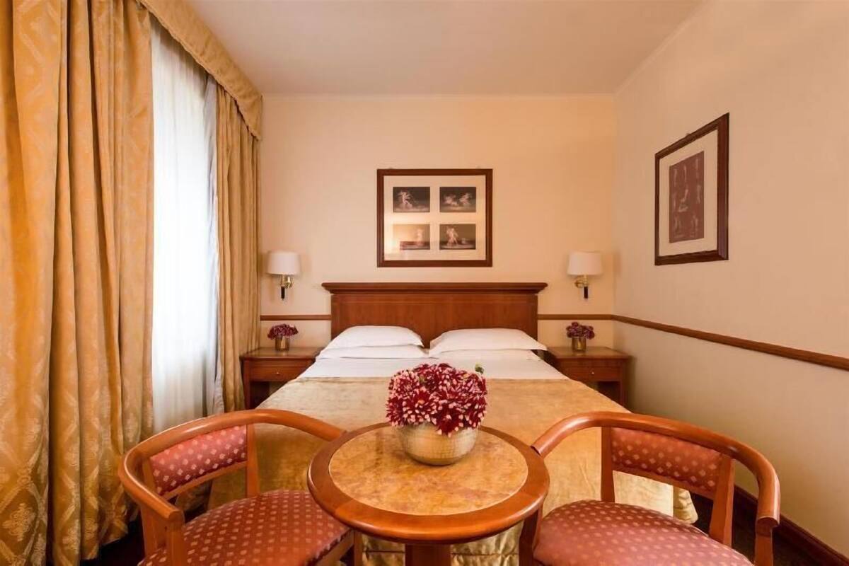 Une chambre d’hôtel comprenant un lit, deux tables de chevet avec des lampes, un tableau encadré au mur, une petite table ronde avec un vase à fleurs et deux chaises.