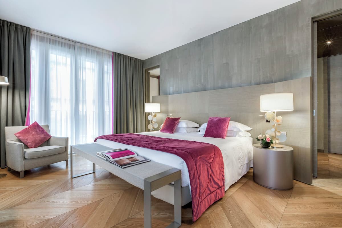 A modern hotel room with a large bed, a bedside table, a chair, and a nightstand with a lamp.