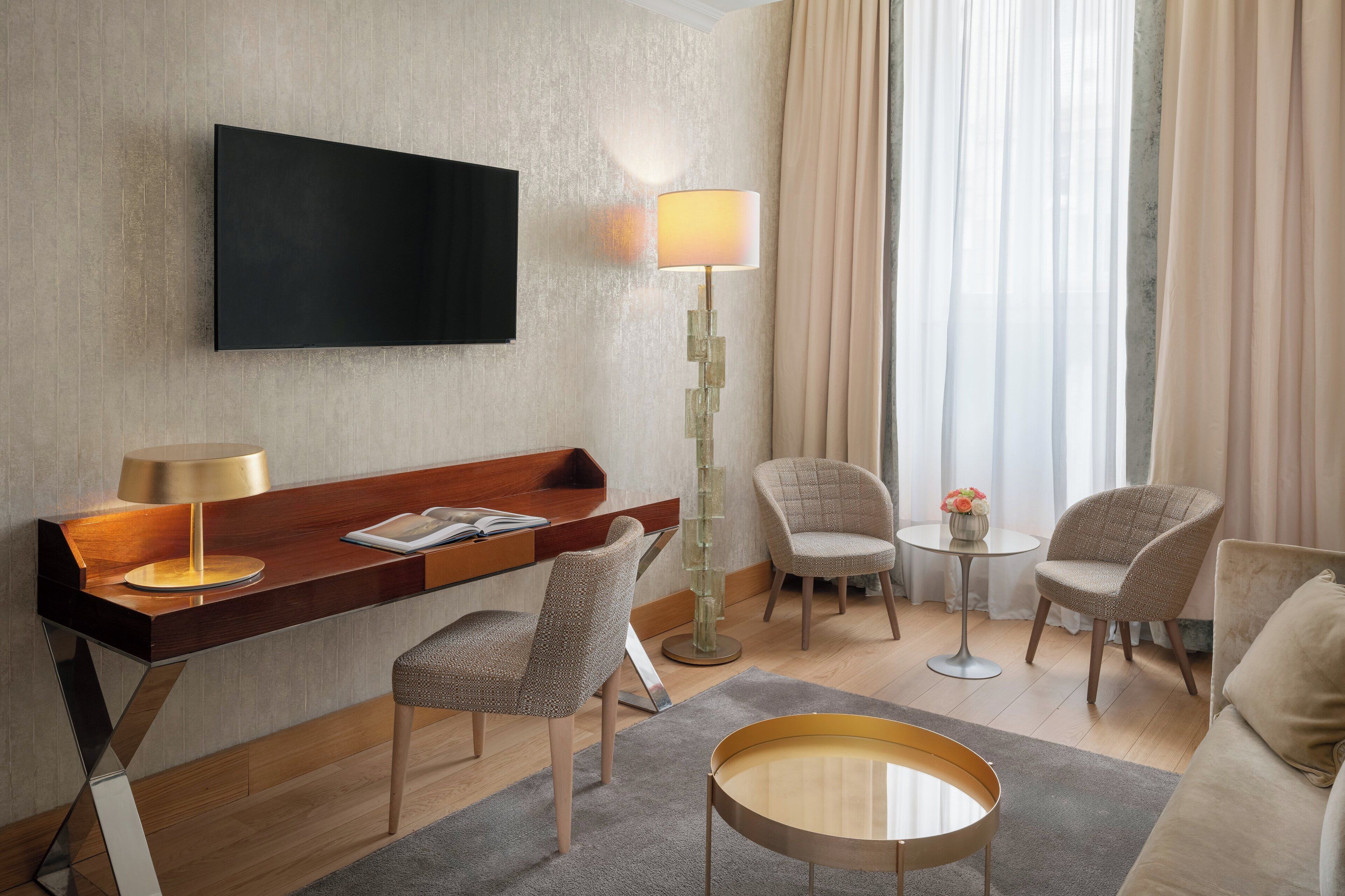 Una habitación de hotel moderna con una televisión de pantalla plana, un escritorio de madera, dos sillones, una pequeña mesa redonda con un florero y una lámpara de pie.