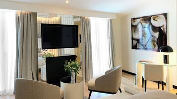 Habitación de hotel moderna con televisión de pantalla plana, sofá, dos sillas, una mesita con una planta y un cuadro en la pared.