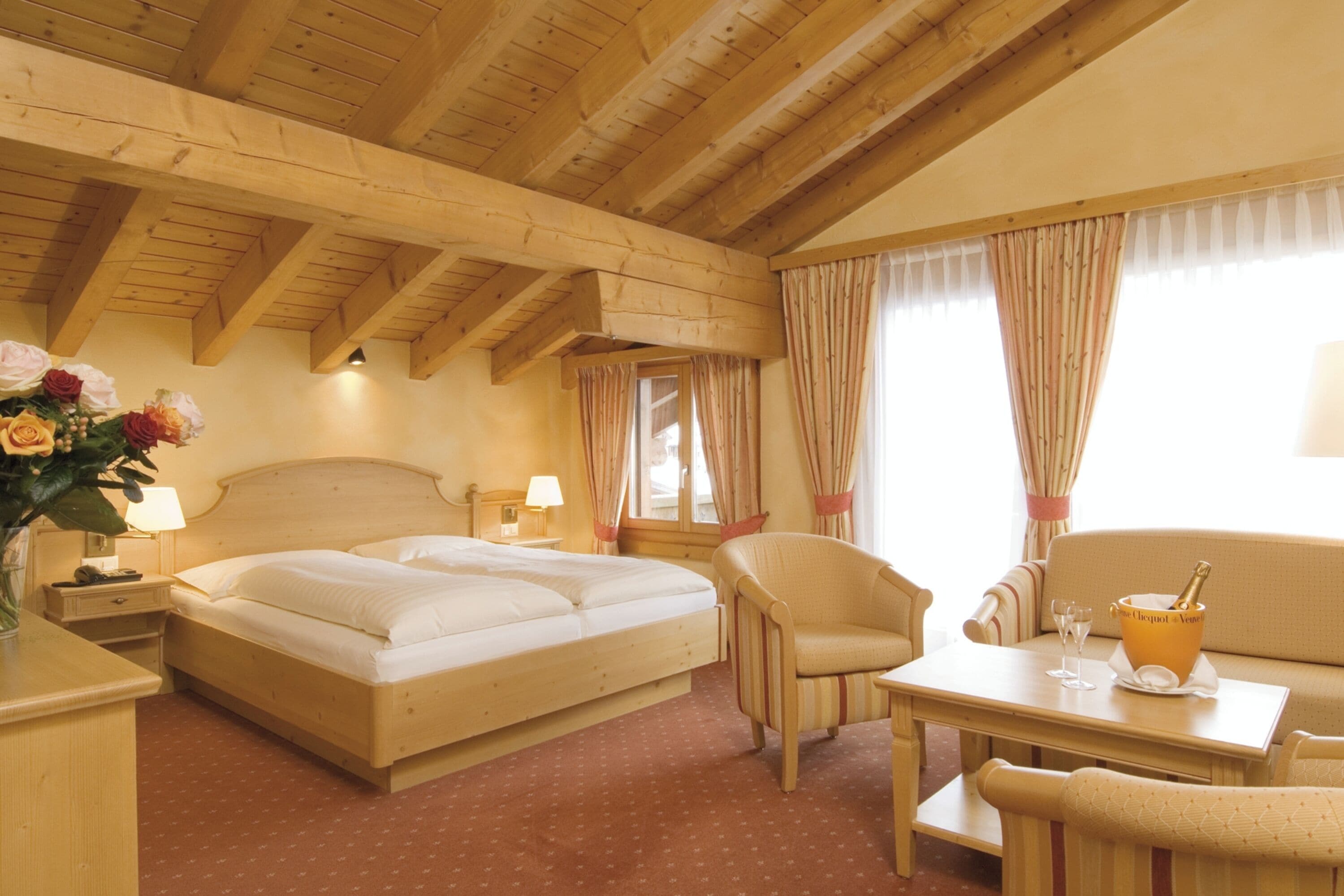 A hotel room with a wooden ceiling, a bed, a chair, a table, and a nightstand with a lamp.