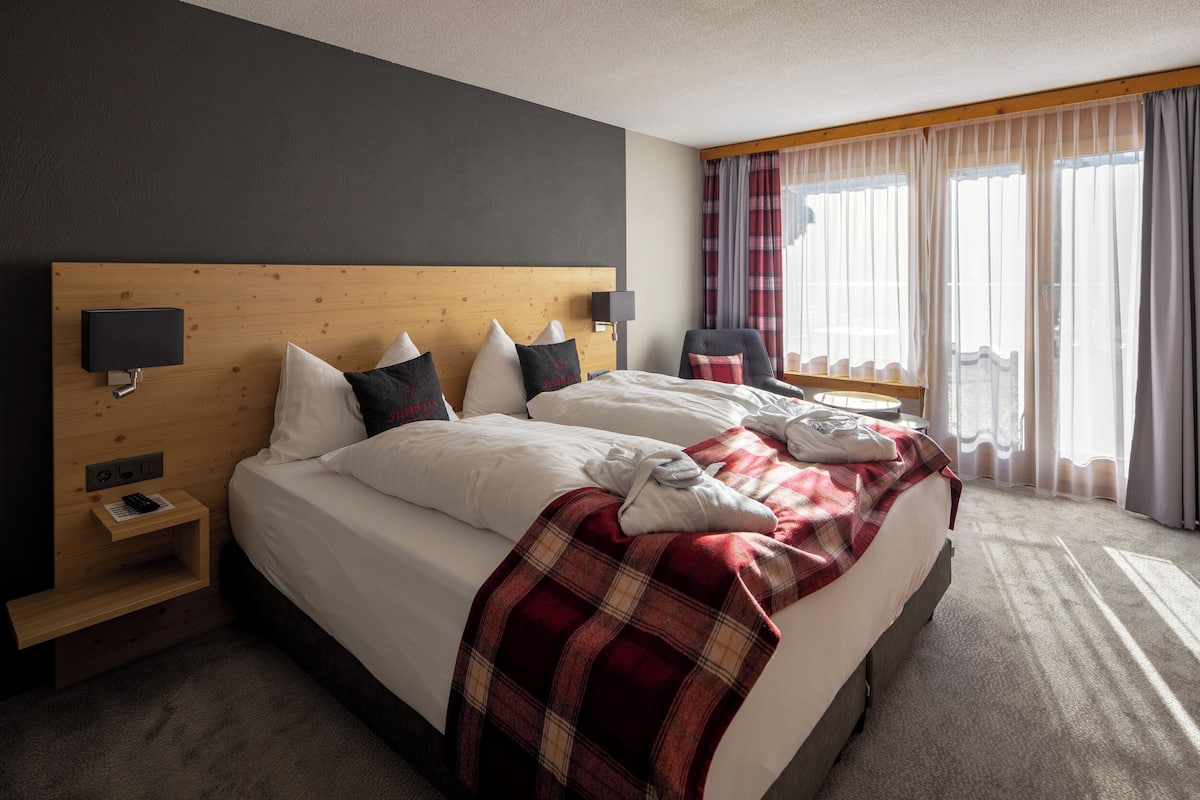 Hotellihuone, jossa on suuri sänky, puinen pääty, yöpöytä lampun kanssa ja ikkuna, jossa on verhot.