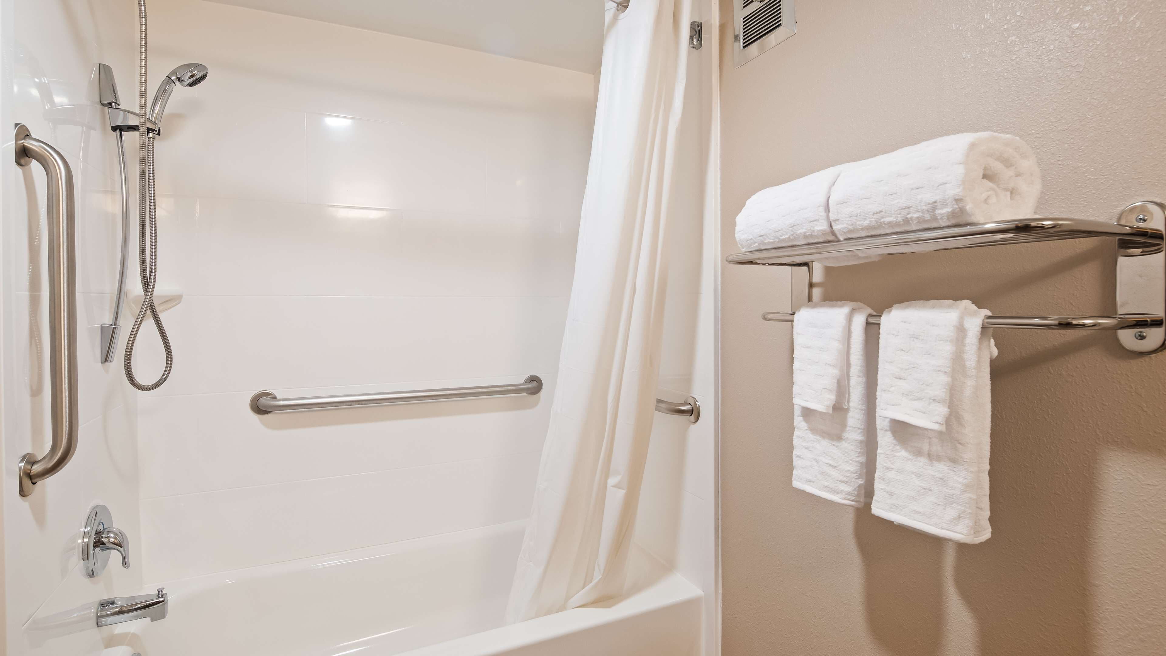 A bathroom with a walk-in shower, grab bars, a towel rack with towels, and a white tiled wall.