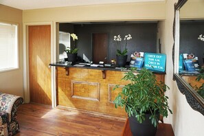 A reception desk with a check-in/out sign, potted plants, and a mirror reflecting the interior.