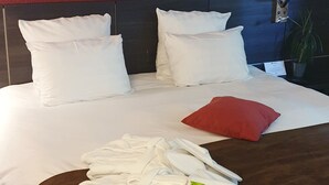 A neatly made bed with white linens and a red pillow, a bedside table with a lamp, and green plants in the foreground. - Alexander Park Chambéry (Chambery)