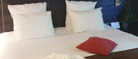 A neatly made bed with white linens and a red pillow, a bedside table with a lamp, and green plants in the foreground.