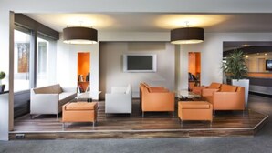 A modern lobby with a seating area featuring orange sofas and white armchairs, a flat-screen TV mounted on the wall, and two pendant lights. - Alexander Park Chambéry (Chambery)
