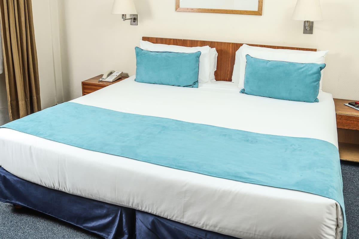 A neatly made bed with a blue bedspread and pillows, a wooden headboard, a bedside table with a lamp, and a phone.
